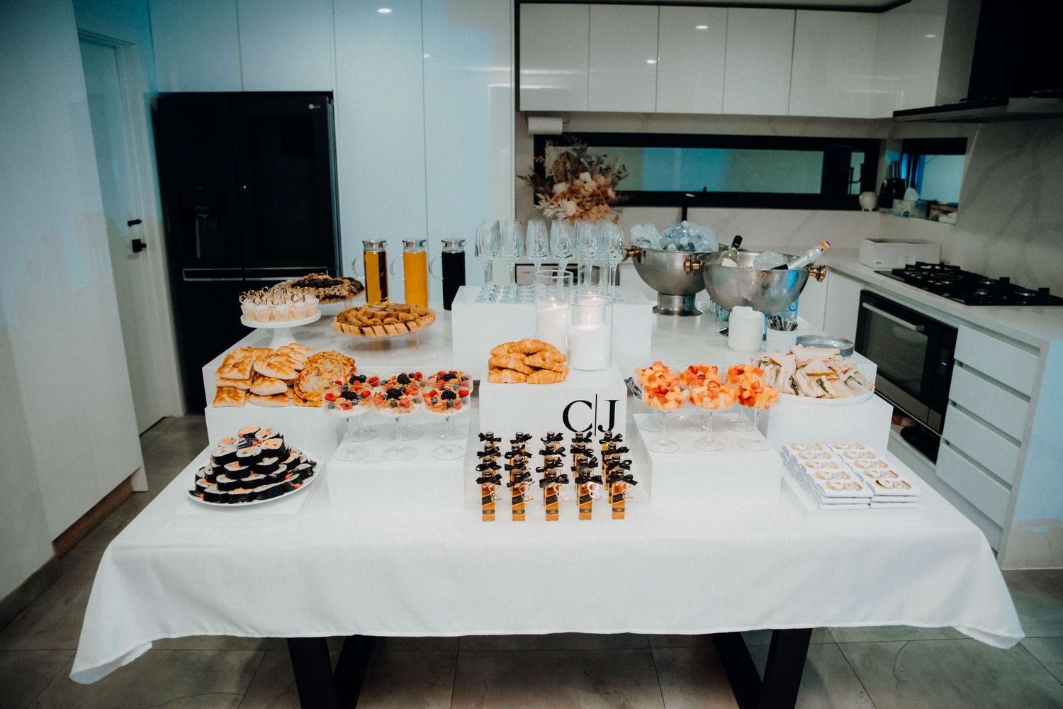 A dessert table with assorted pastries, chocolates, and beverages displayed on a white tablecloth in a modern kitchen.