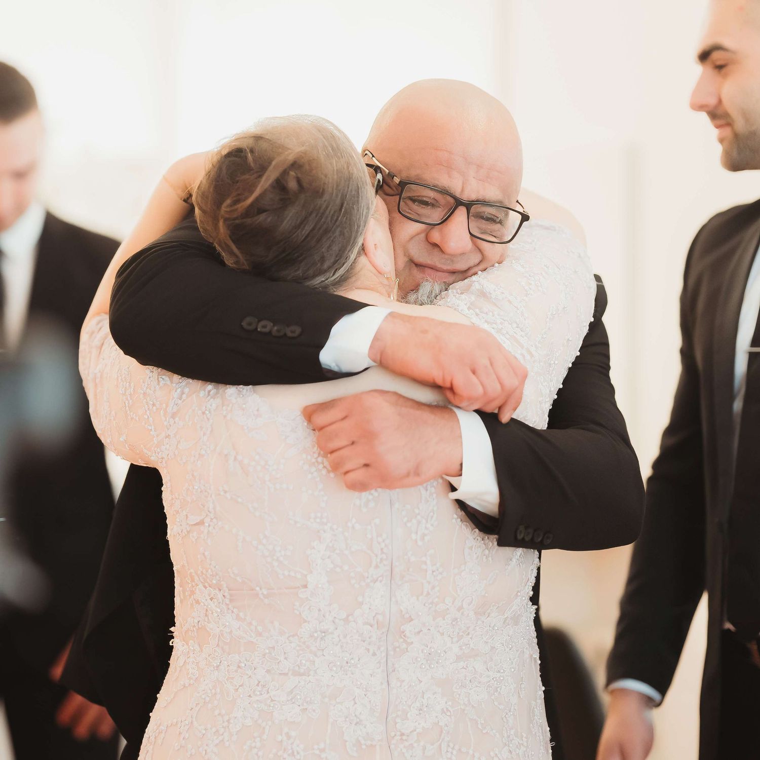 Two people share an emotional embrace at what appears to be a wedding ceremony while others look on in the background.