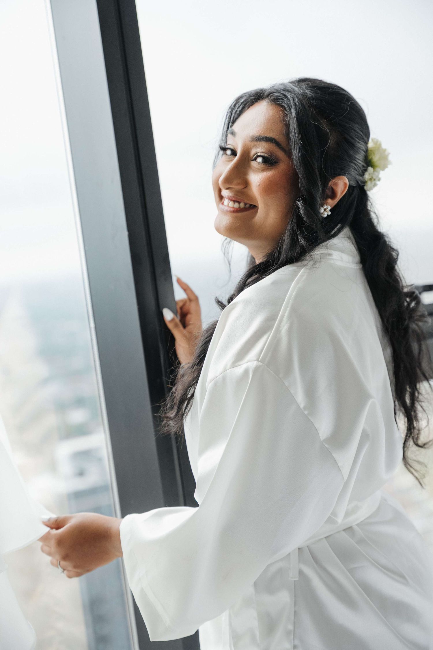 Smiling woman in white robe stands by window overlooking city view, with flower in her dark braided hair.