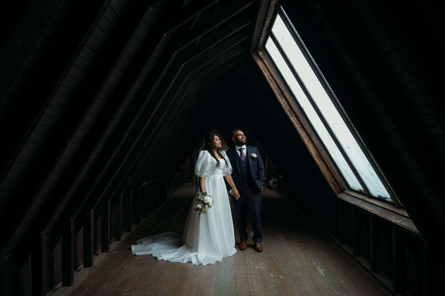 A bride and groom sit together in a dark, dramatic attic space with light streaming through a skylight window.