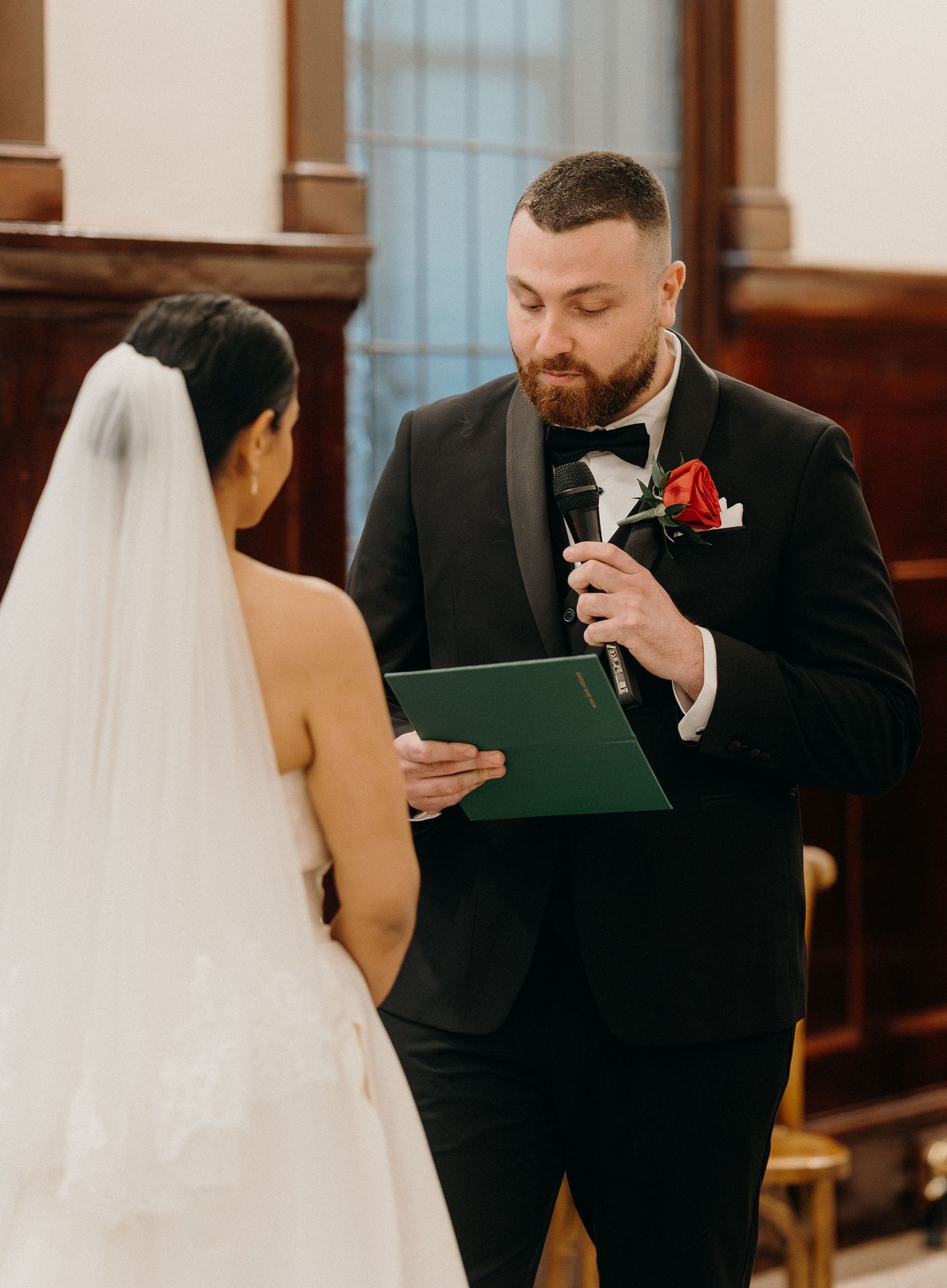 Groom in black tuxedo reads wedding vows from green folder to bride in white gown during ceremony.
