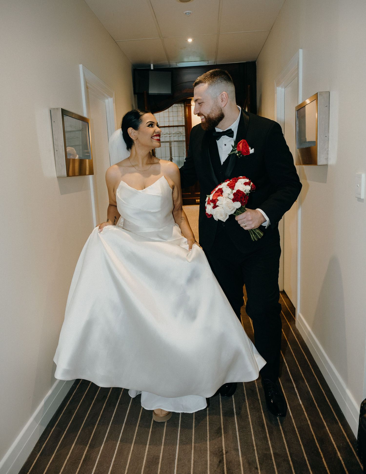 Bride in white strapless gown and groom in black tuxedo walk together in a hallway holding red and white bouquet.