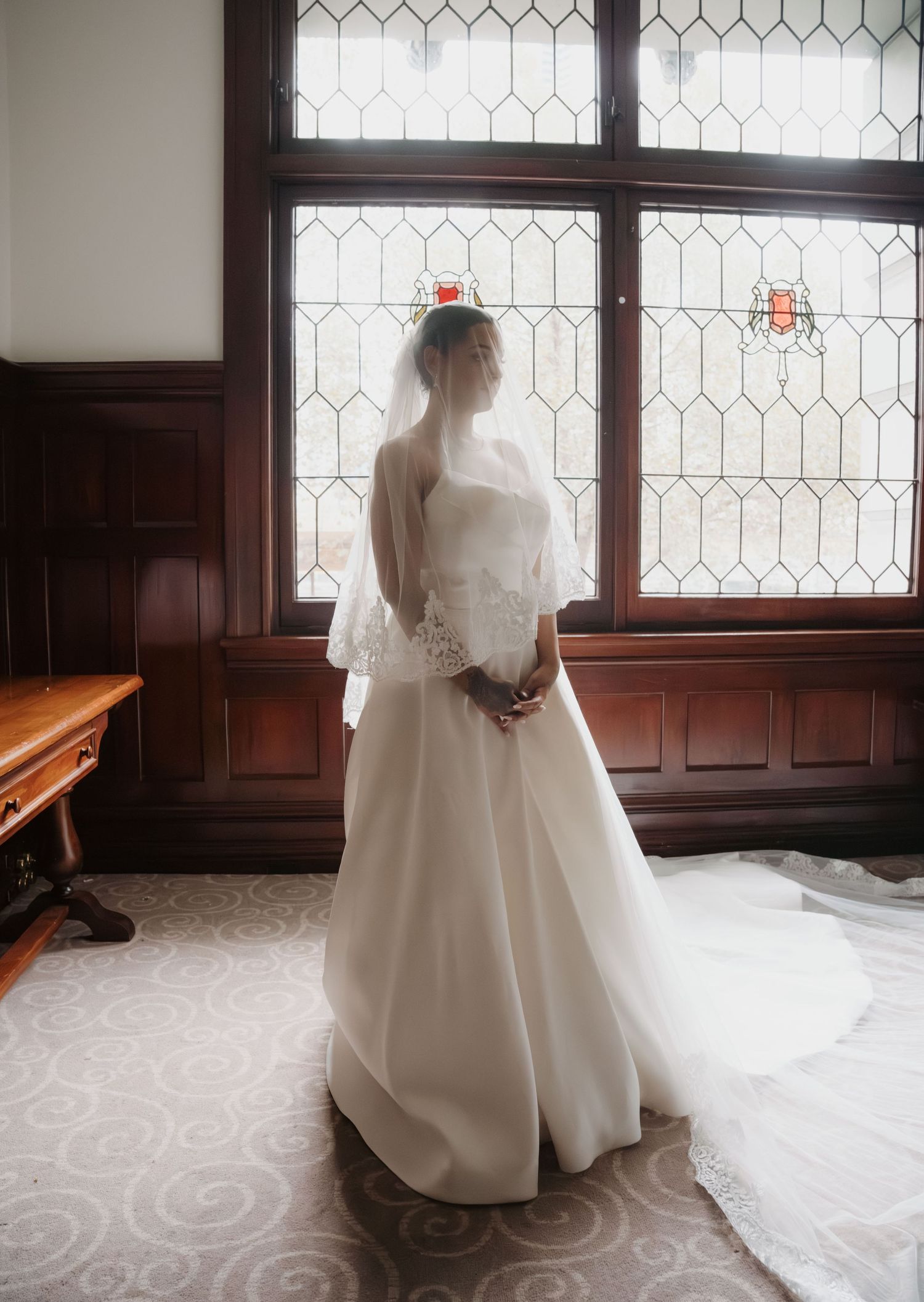 Bride in white wedding gown and veil stands before ornate leaded glass windows in elegant wood-paneled room.