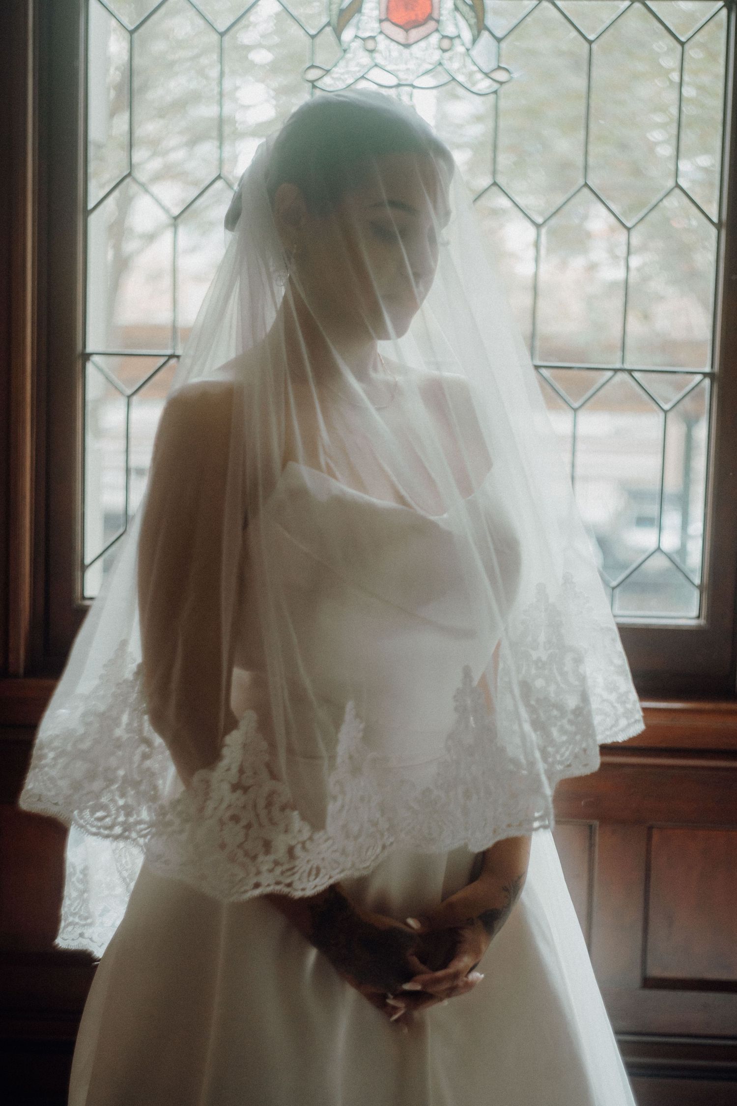 Bride in a white strapless gown and lace-trimmed veil stands before a stained glass window in soft natural light.