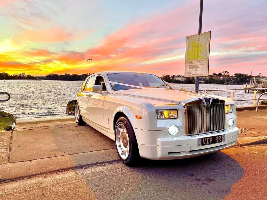 A luxurious white Rolls-Royce stretch limousine parked on a beach at sunset with pink and orange skies.