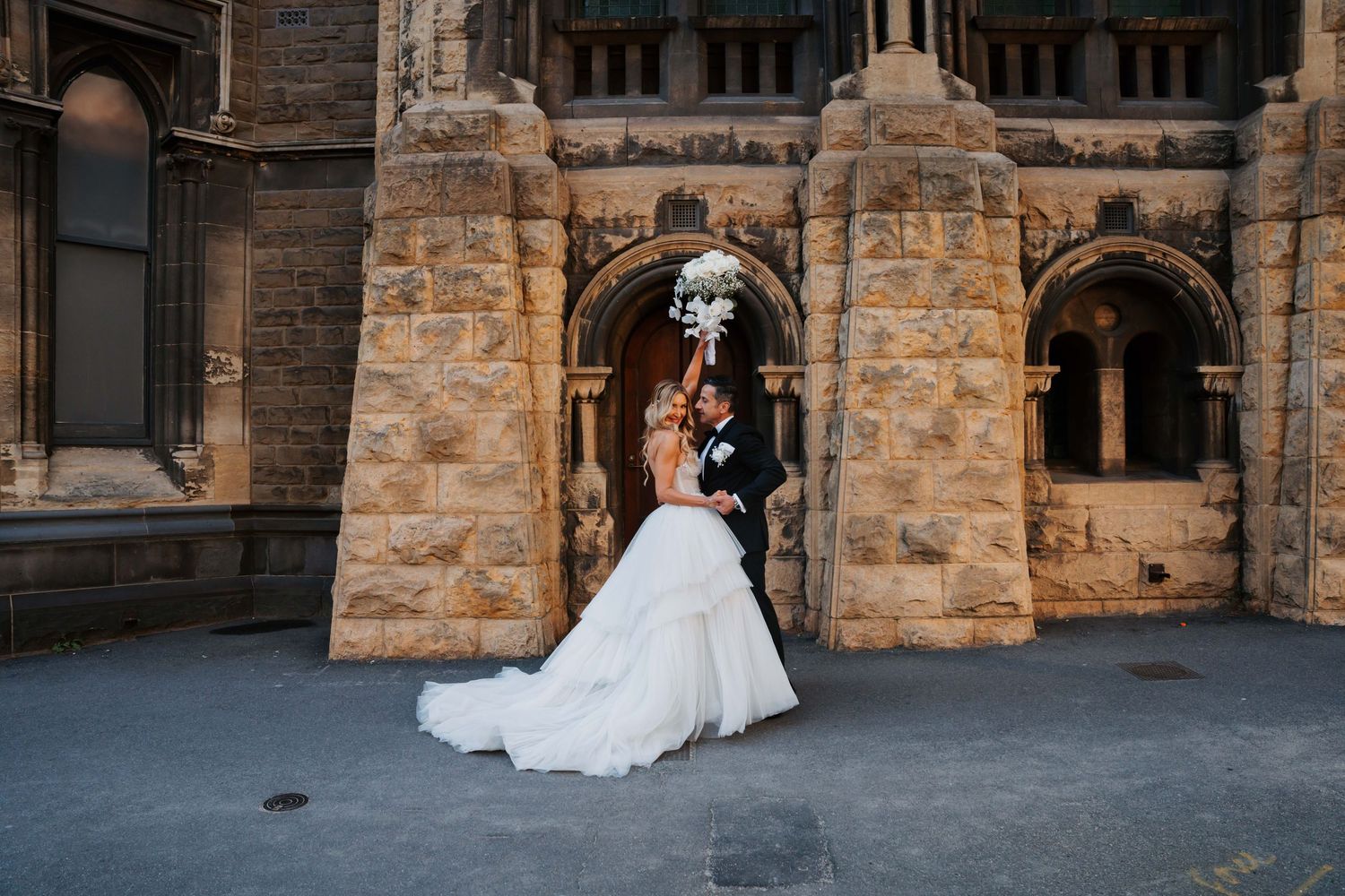 A wedding couple embraces in front of a historic stone church building with arched architectural details.