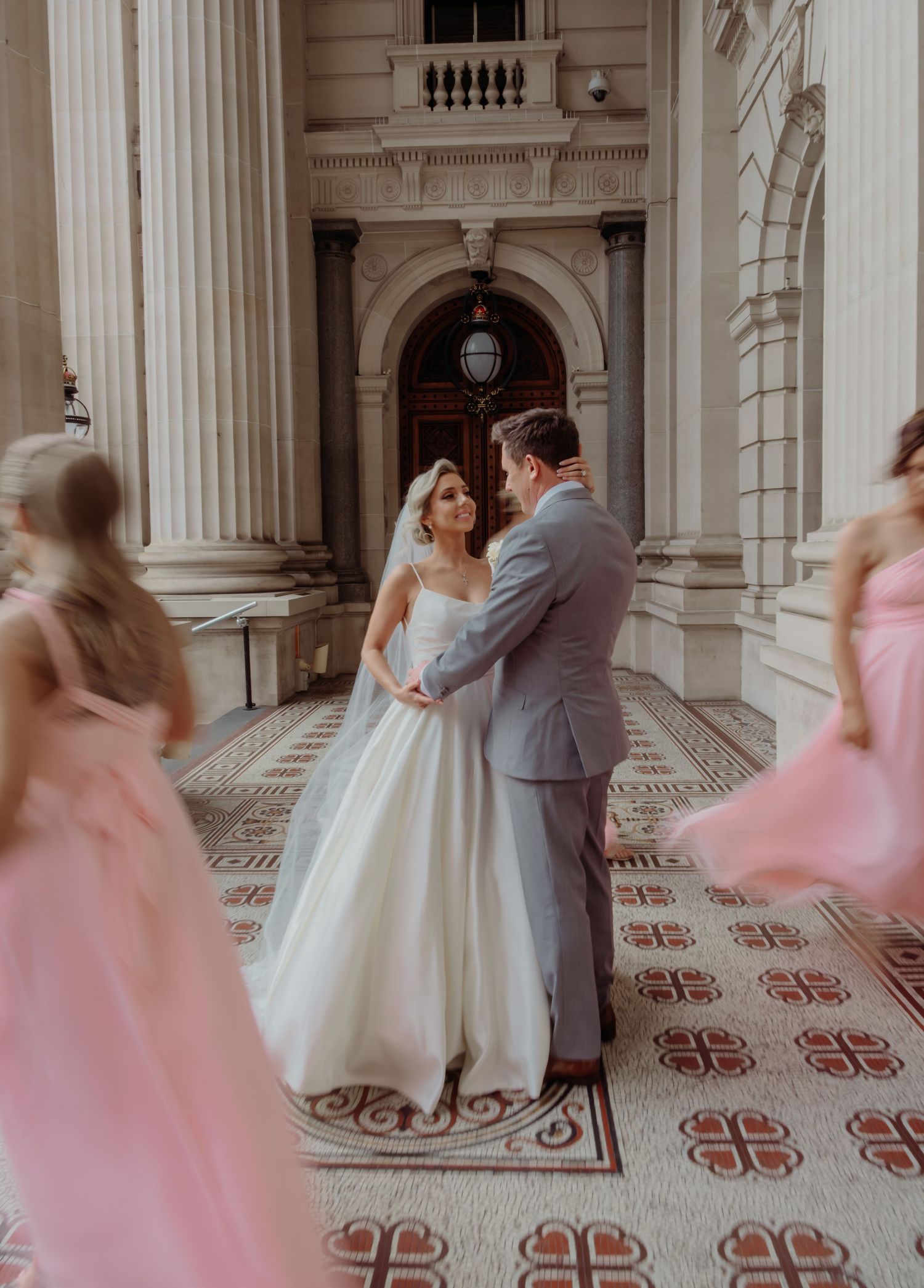 A couple shares an intimate moment on ornate tiled floors surrounded by classical architecture and pink-dressed figures in motion.