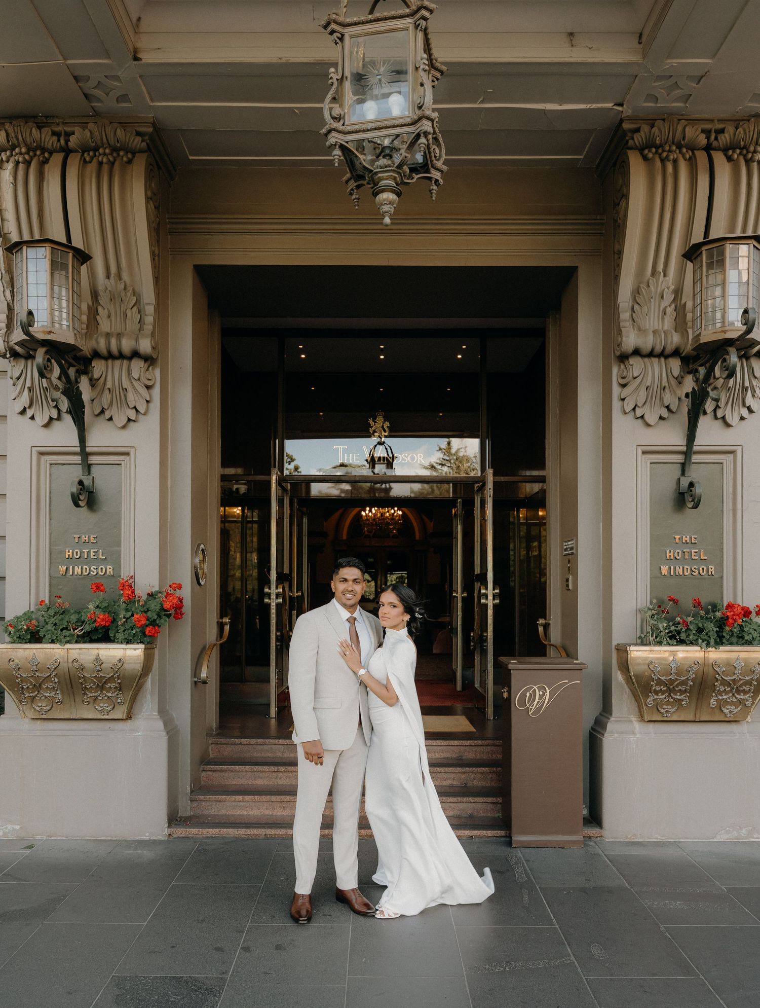 A couple in wedding attire poses elegantly in front of a grand hotel entrance with ornate architecture.