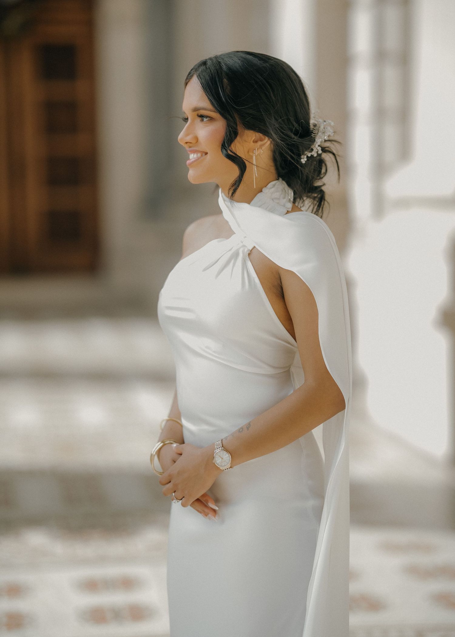 A woman in an elegant white one-shoulder gown poses gracefully in a bright sunlit hallway with wooden accents.