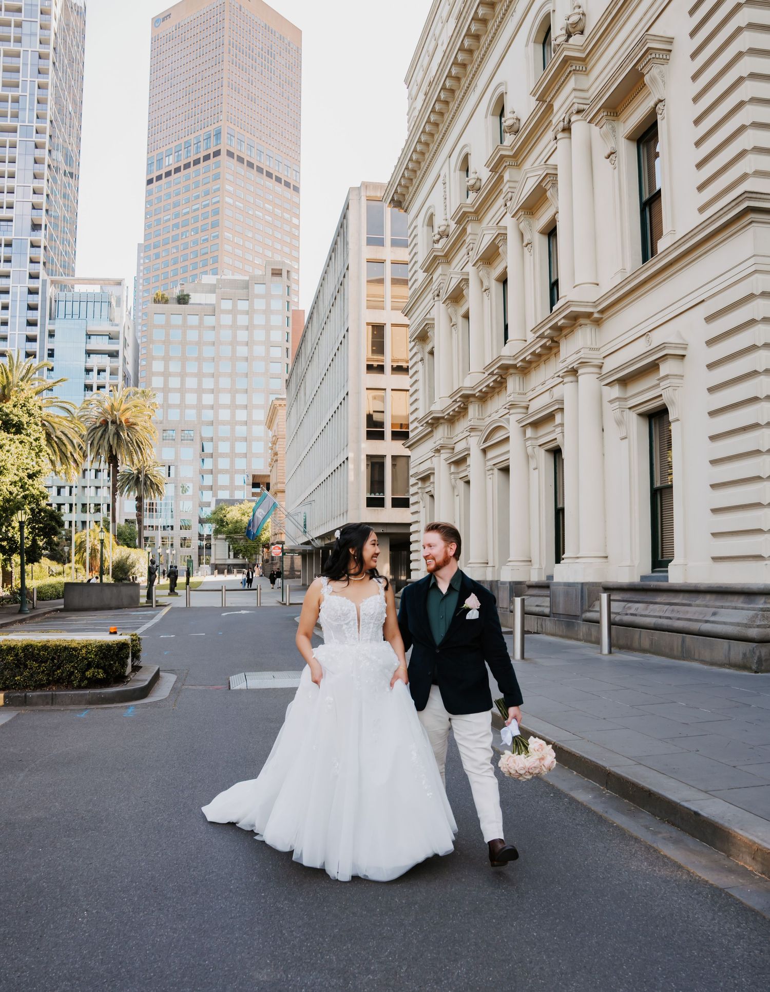 A couple in wedding attire walk together outside an ornate historic building in an urban setting with modern skyscrapers behind them.