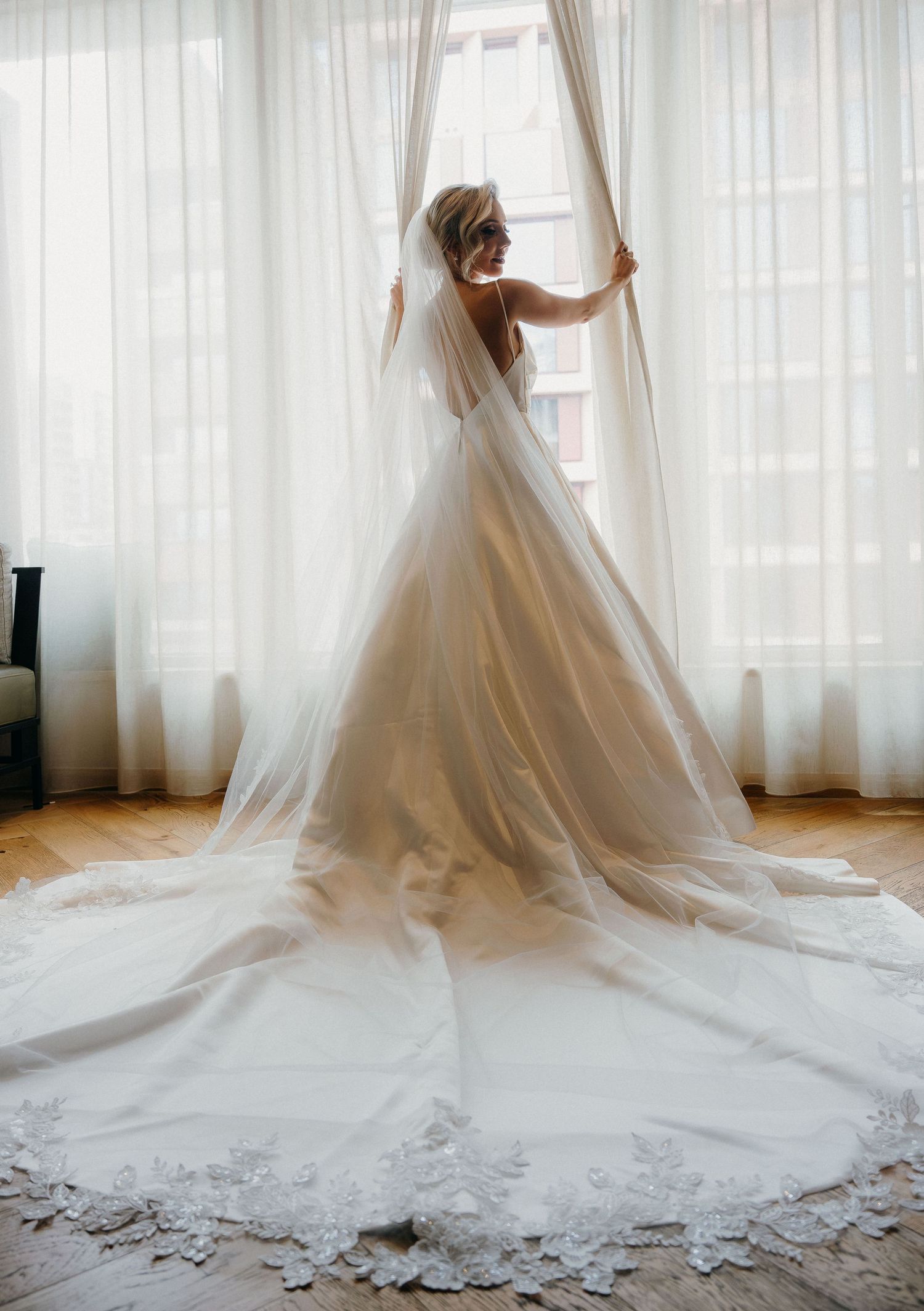 A stunning white wedding gown with a flowing cathedral train is displayed against sheer curtains in a bright room.