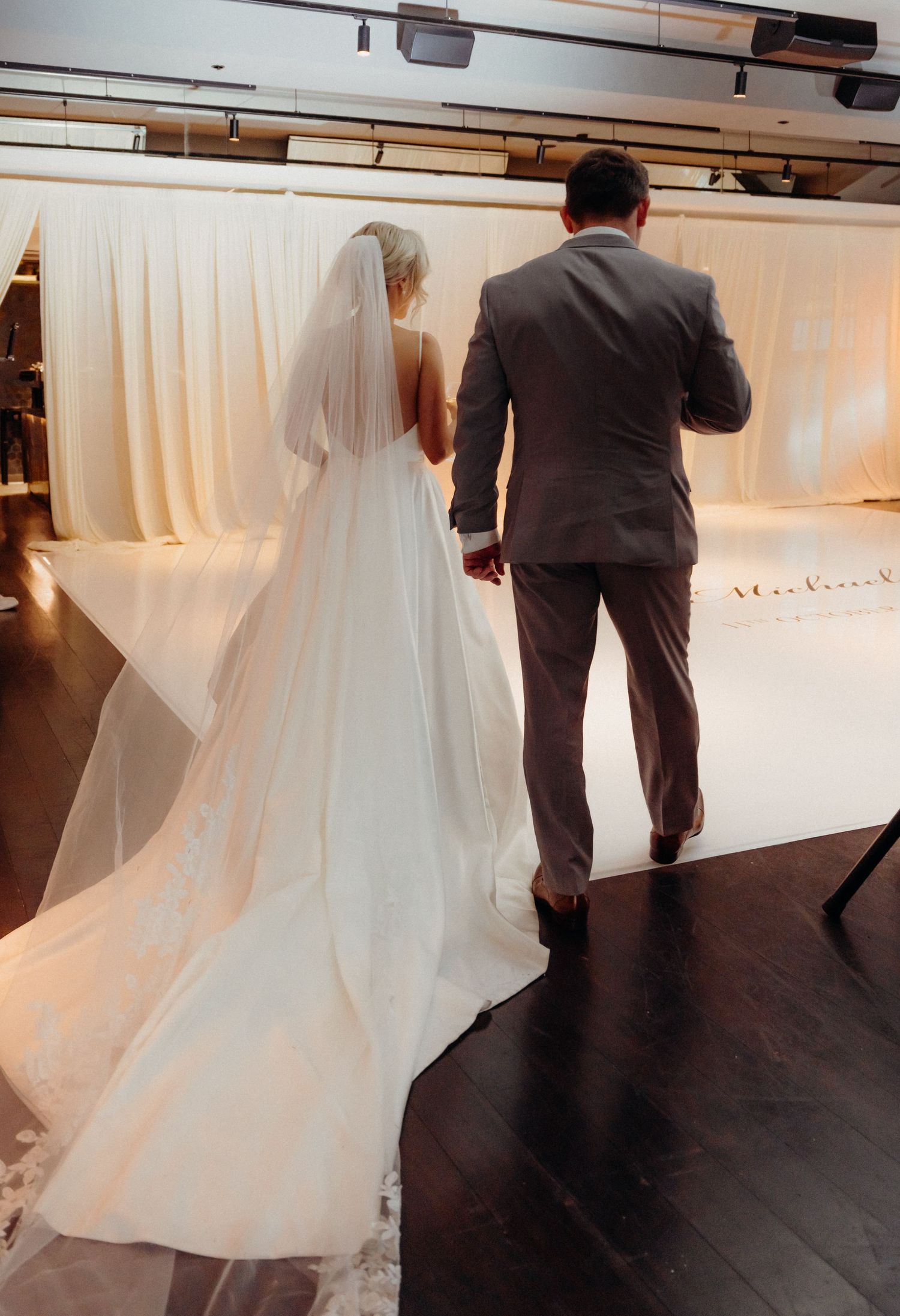 A couple in formal wedding attire dances together on a dark wooden floor during their first dance at a reception.