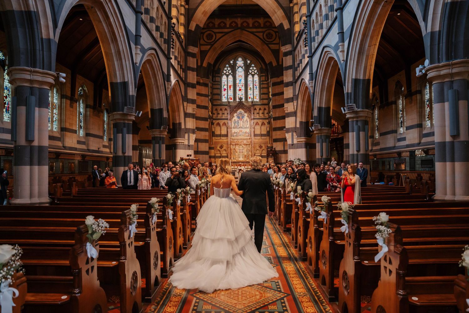 Ornate church interior with wooden pews and stained glass windows illuminated by warm lighting during a ceremony.