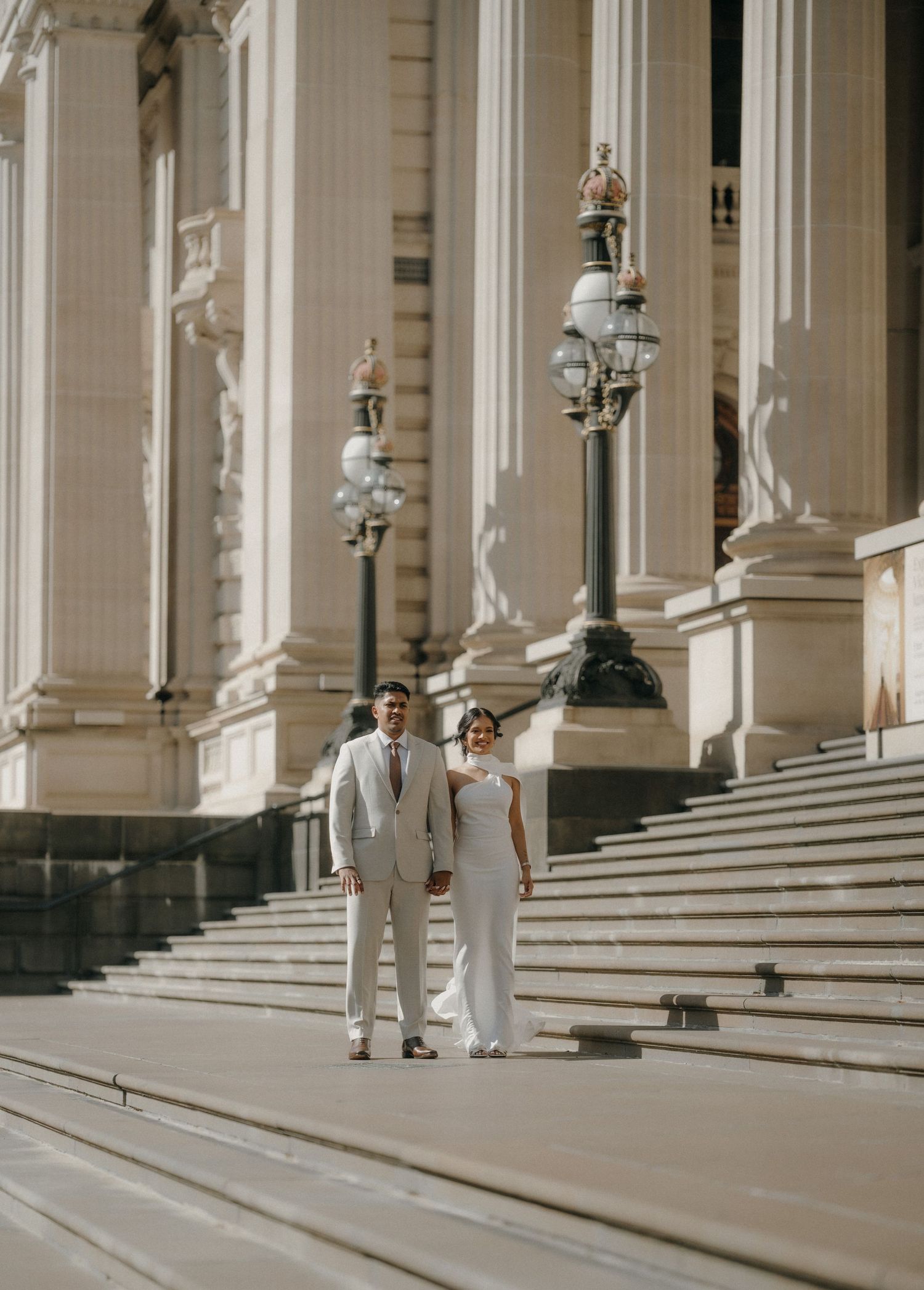 A couple in elegant attire poses on grand marble steps of a classic architectural building in warm sunlight.