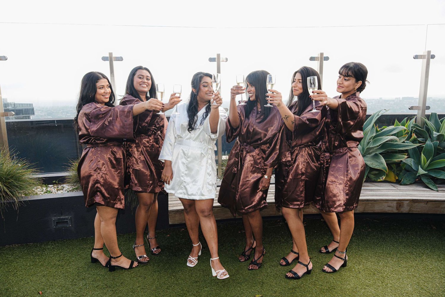 A wedding party in matching brown satin robes poses together on an outdoor rooftop garden terrace.