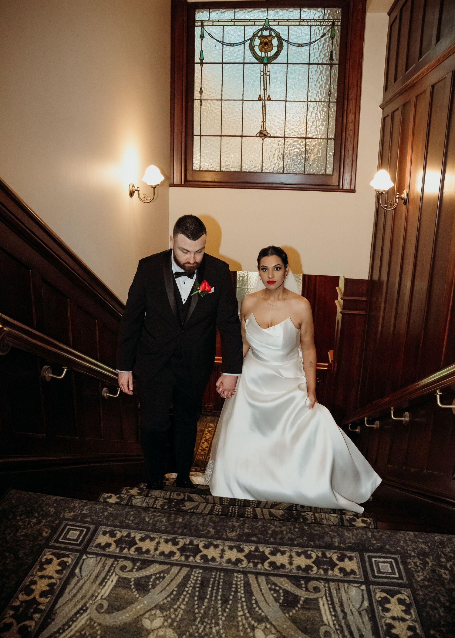 Bride and groom descend grand staircase, she in white satin gown, he in black tuxedo, beneath stained glass window.