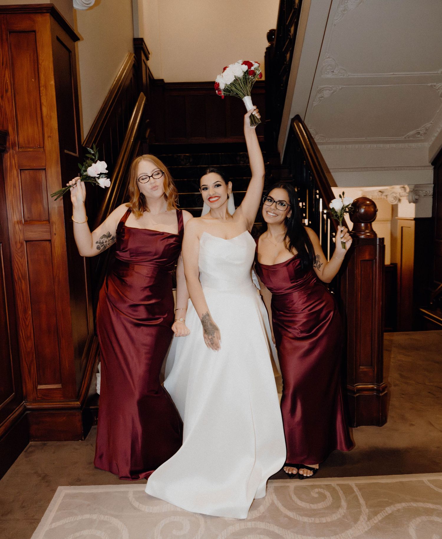 Bride in white gown celebrates with two bridesmaids in burgundy satin dresses on elegant staircase.