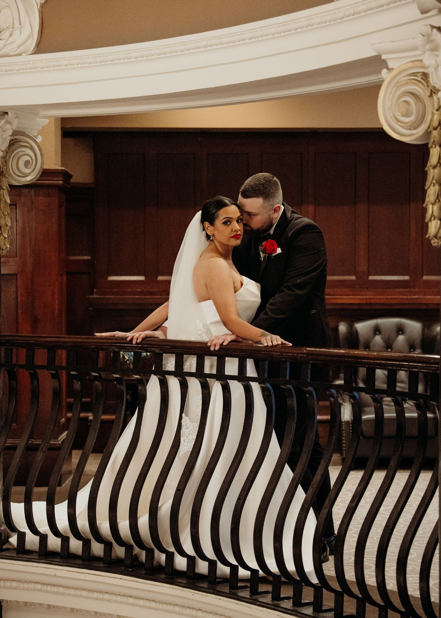 Bride and groom posing on elegant staircase balcony with striped railing, bride in white gown with veil, groom in black suit.