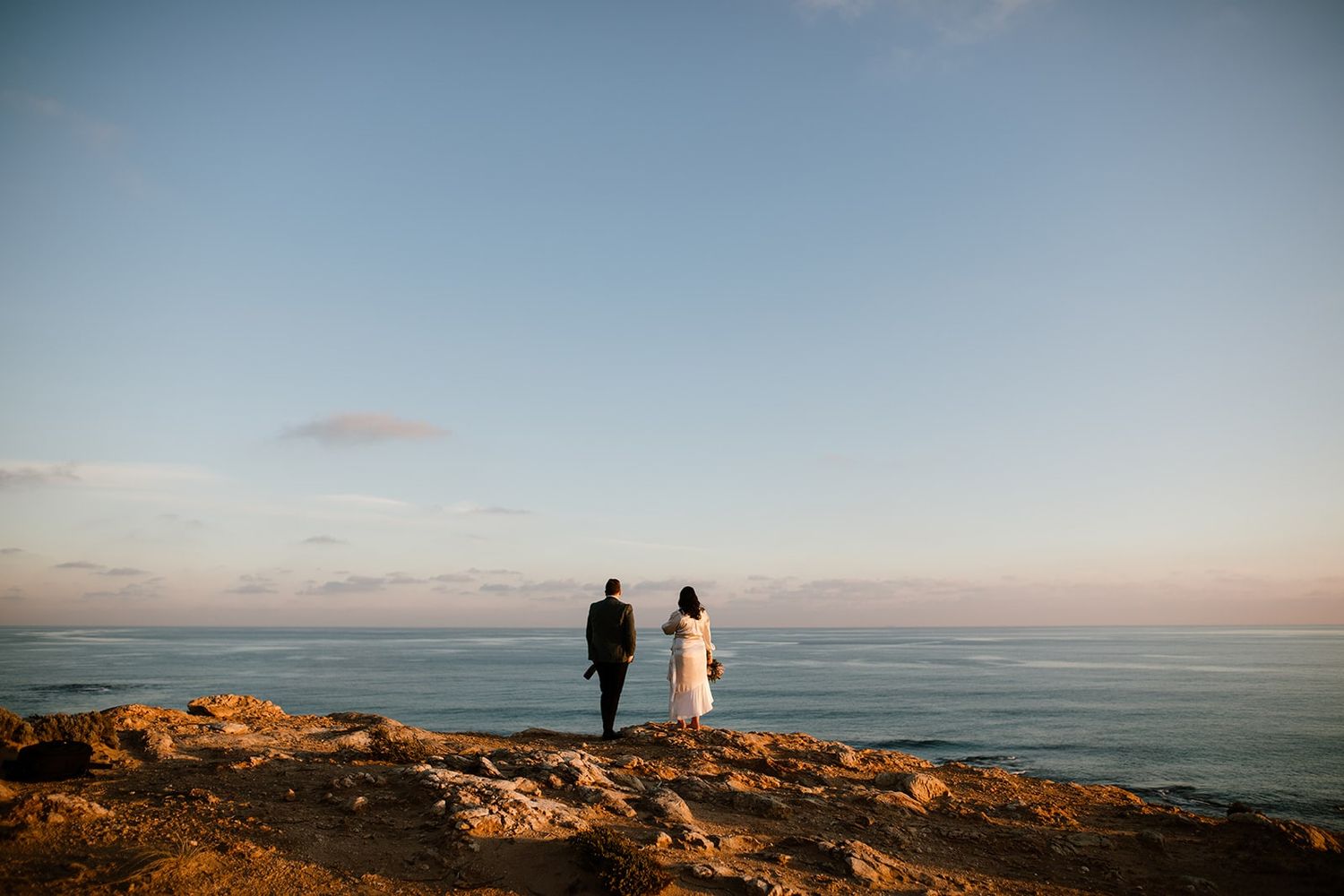 Two silhouettes stand on a coastal cliff against a serene ocean horizon during golden hour.