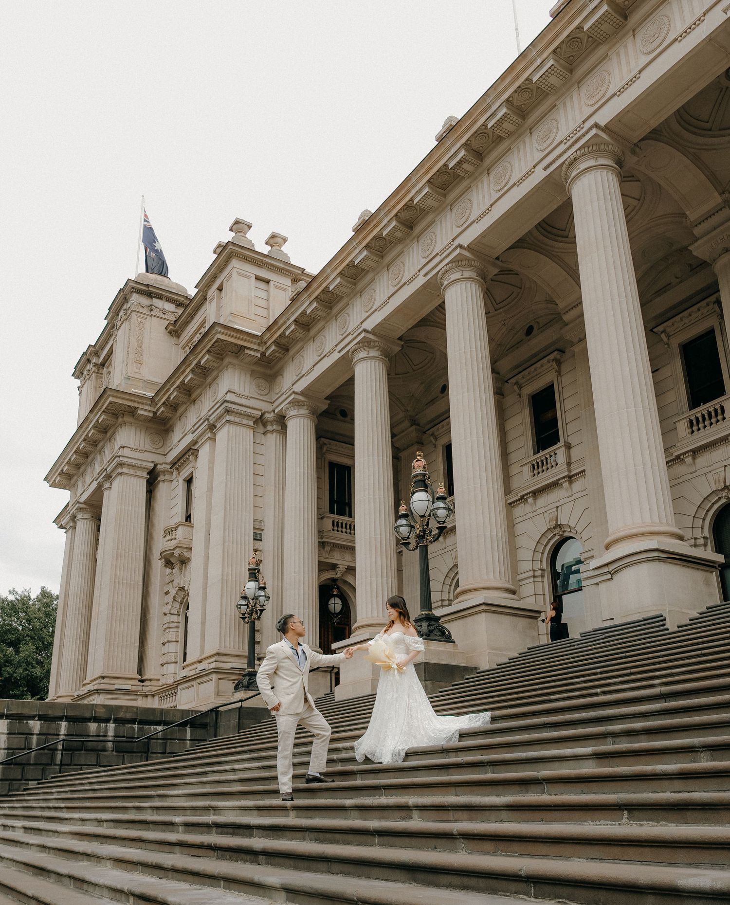 A wedding couple descends the grand marble staircase of a historic building with white neoclassical columns.