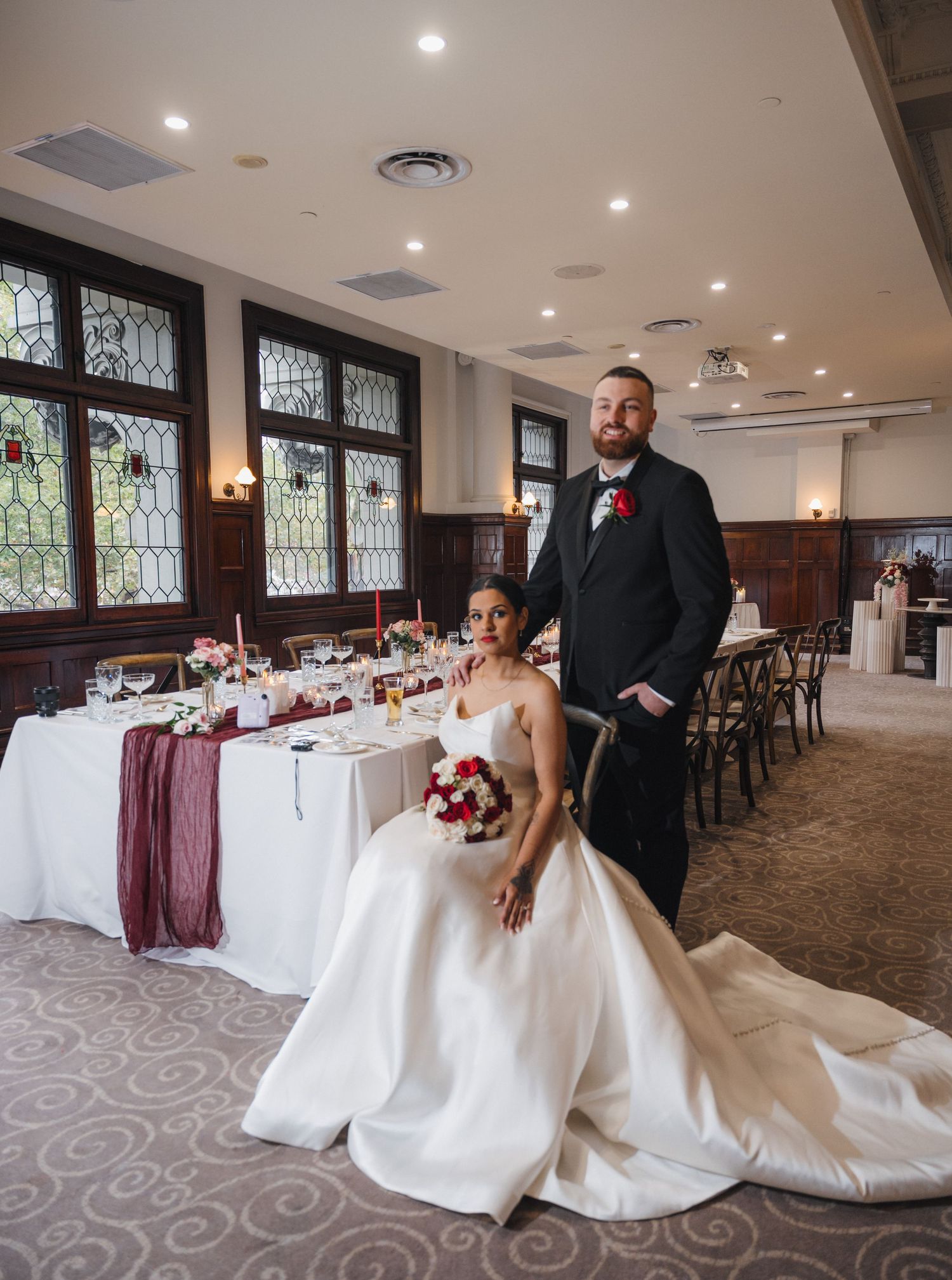 Bride in white gown and groom in black suit pose at decorated wedding reception table with burgundy accents.