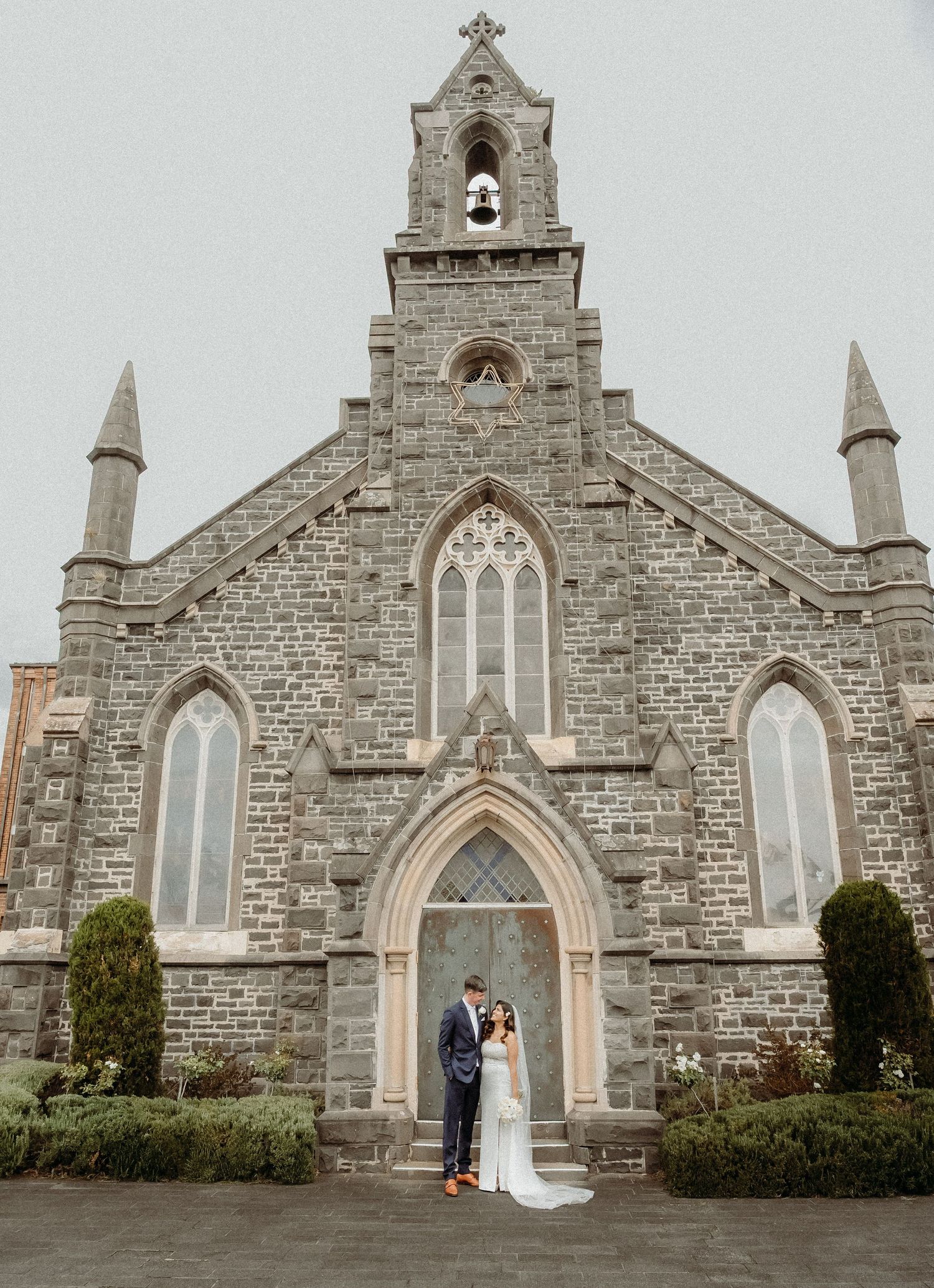 A couple stands in front of a stone Gothic church facade with arched windows and ornate architectural details.