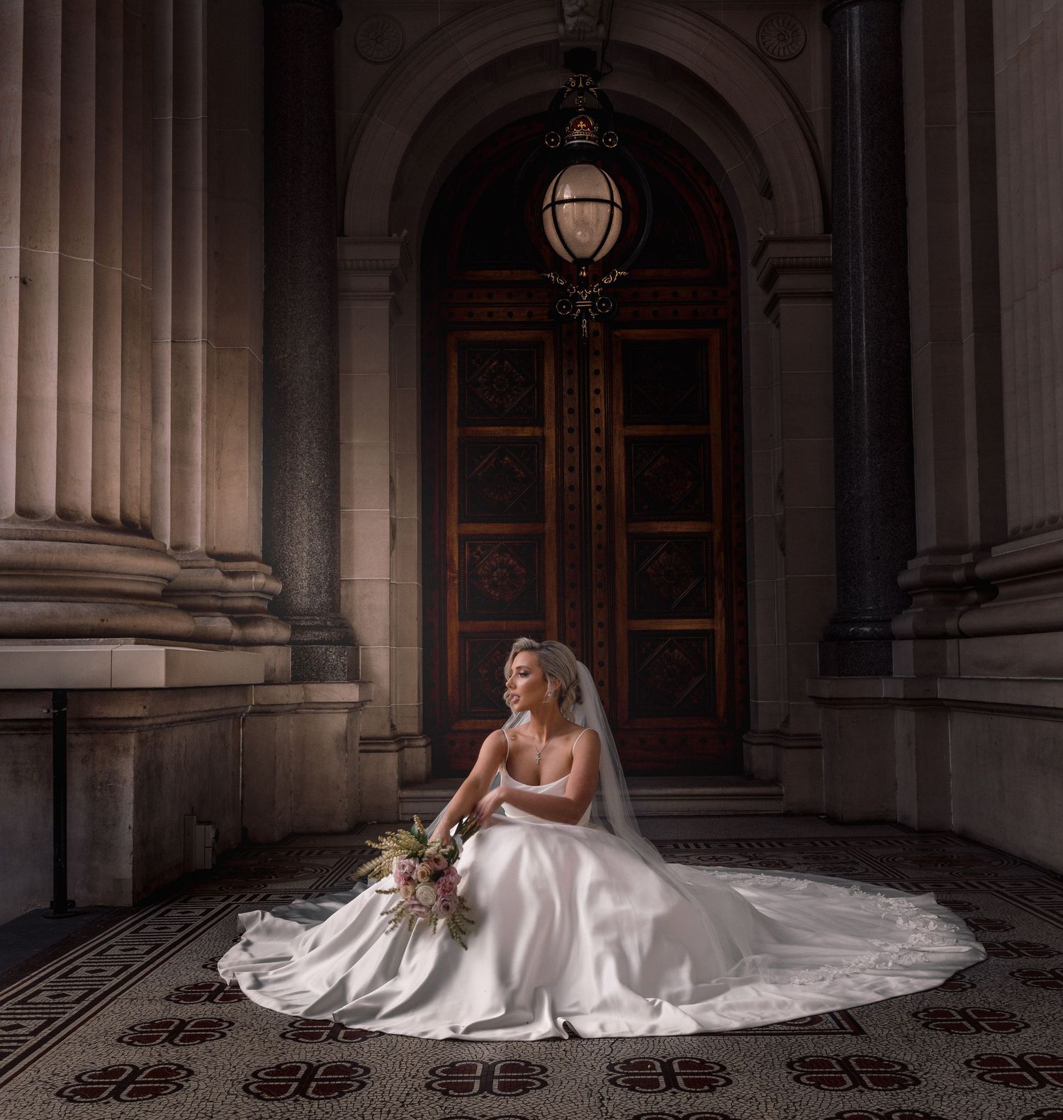 A bride in a flowing white dress sits gracefully on ornate stone steps in a grand architectural setting.