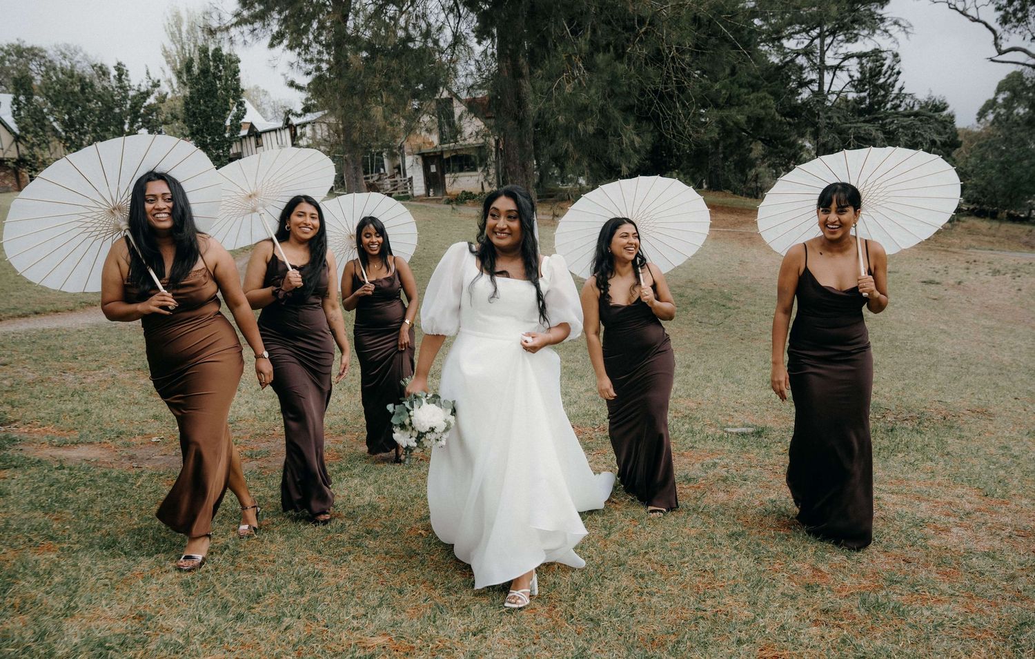 Wedding party in black dresses and white parasols pose playfully in a grassy outdoor setting.