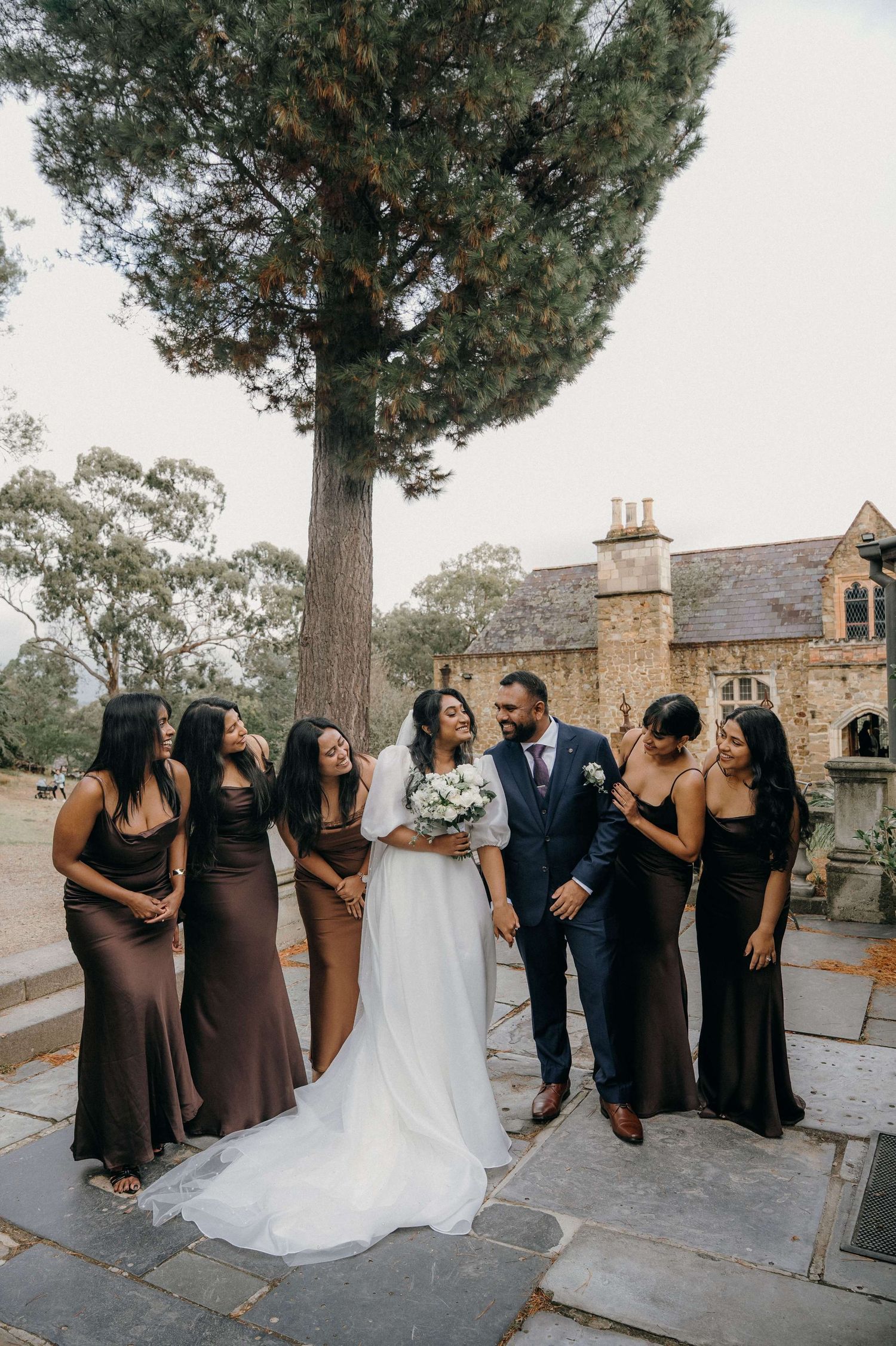 Wedding party poses together on stone patio with tall pine tree and stone building in background.