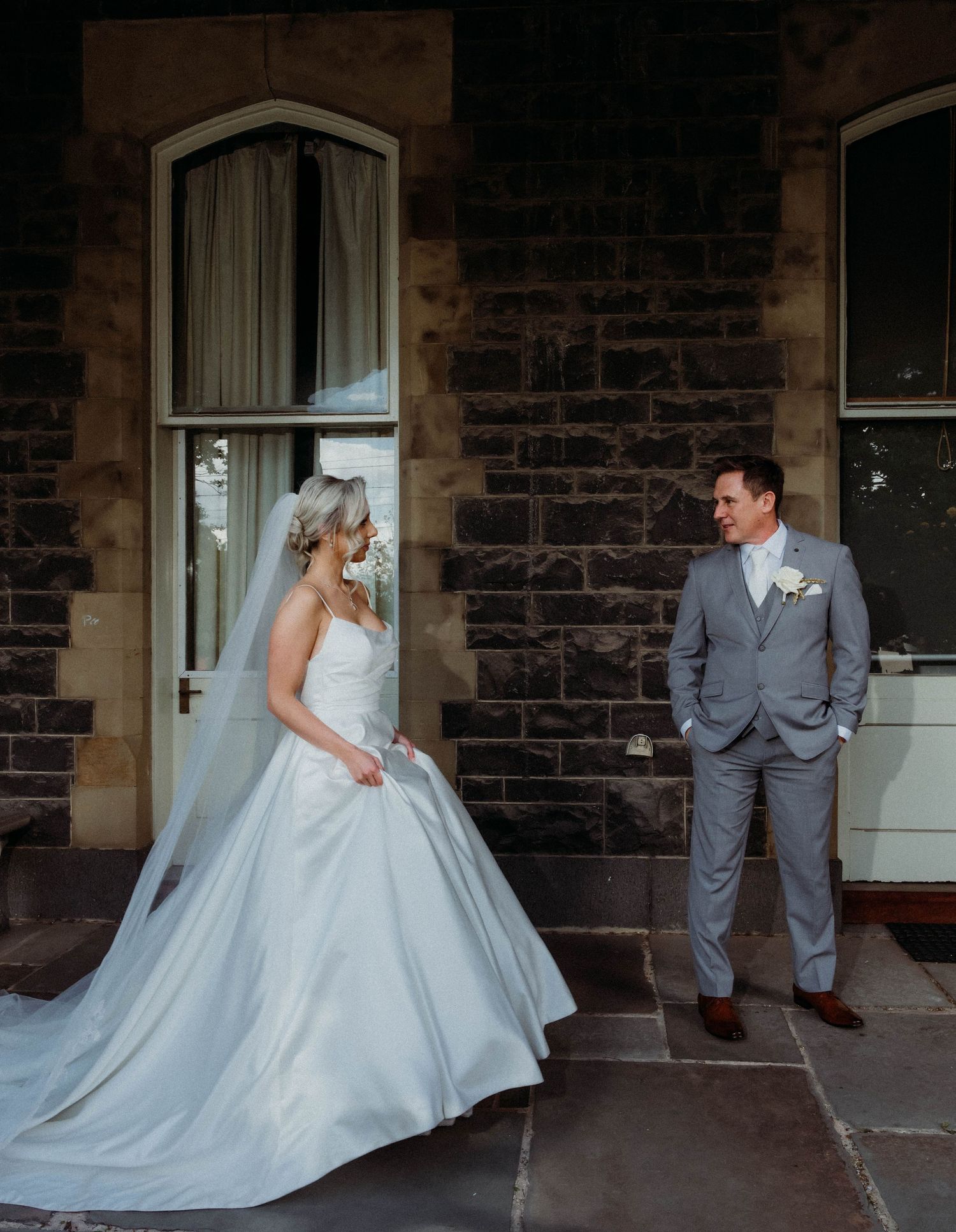 A bride in a flowing white gown and groom in grey suit share playful moments on a stone porch during their wedding shoot.