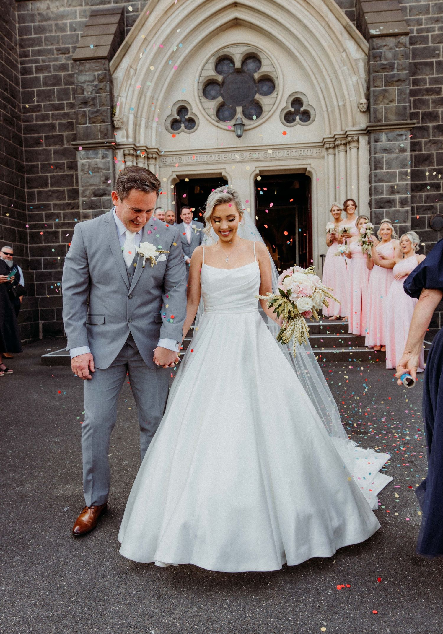A wedding couple exits a gothic church entrance while bridesmaids in pink dresses look on as confetti falls around them.