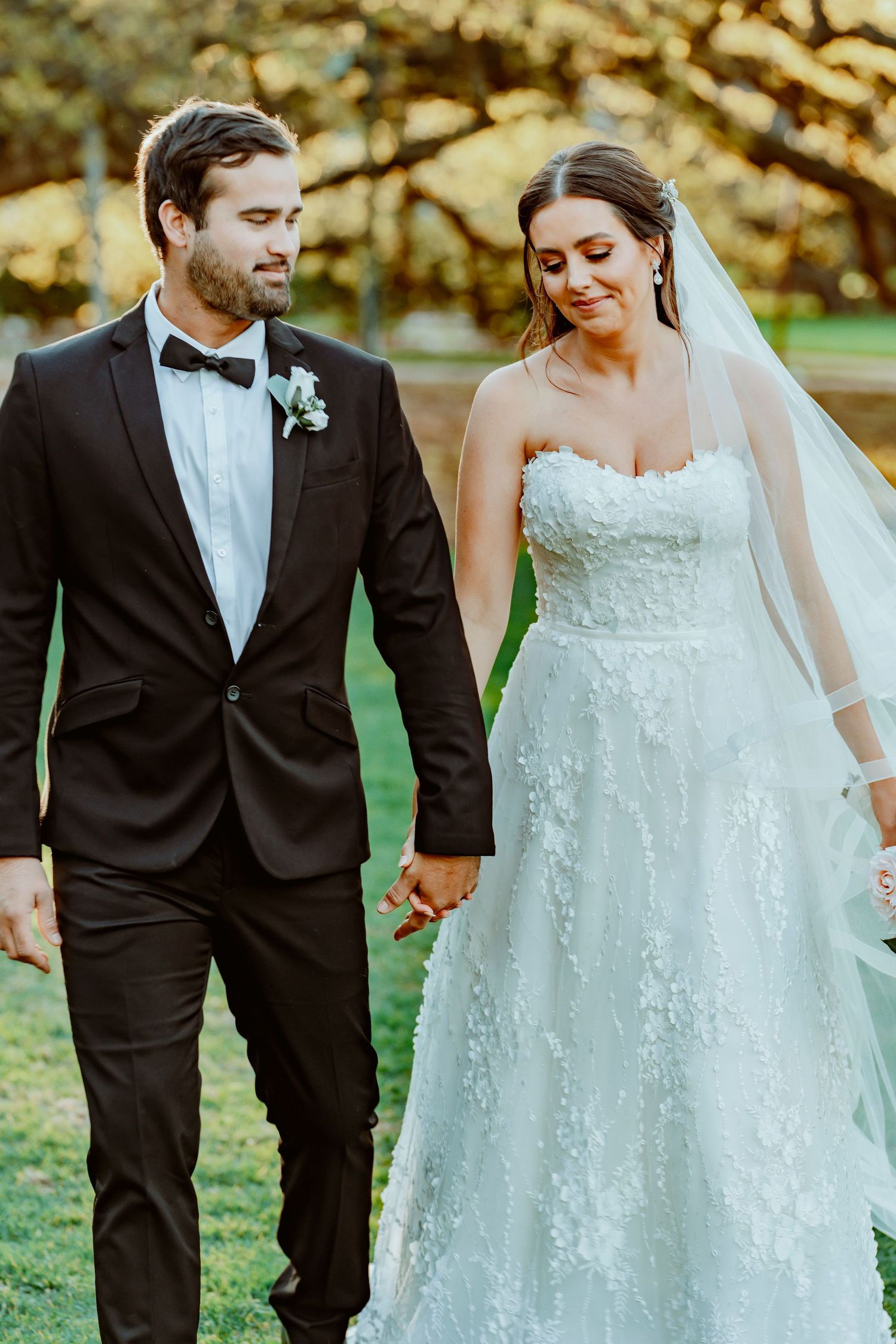 Newlyweds walking hand in hand outdoors, with bride in lace gown and groom in classic black tuxedo at sunset.