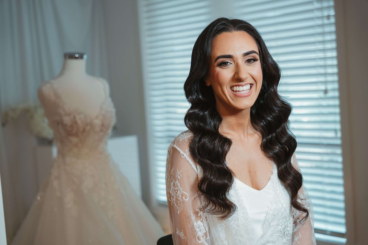 A bride getting ready on her wedding day surrounded by multiple hanging white gowns and dresses in a bridal suite.