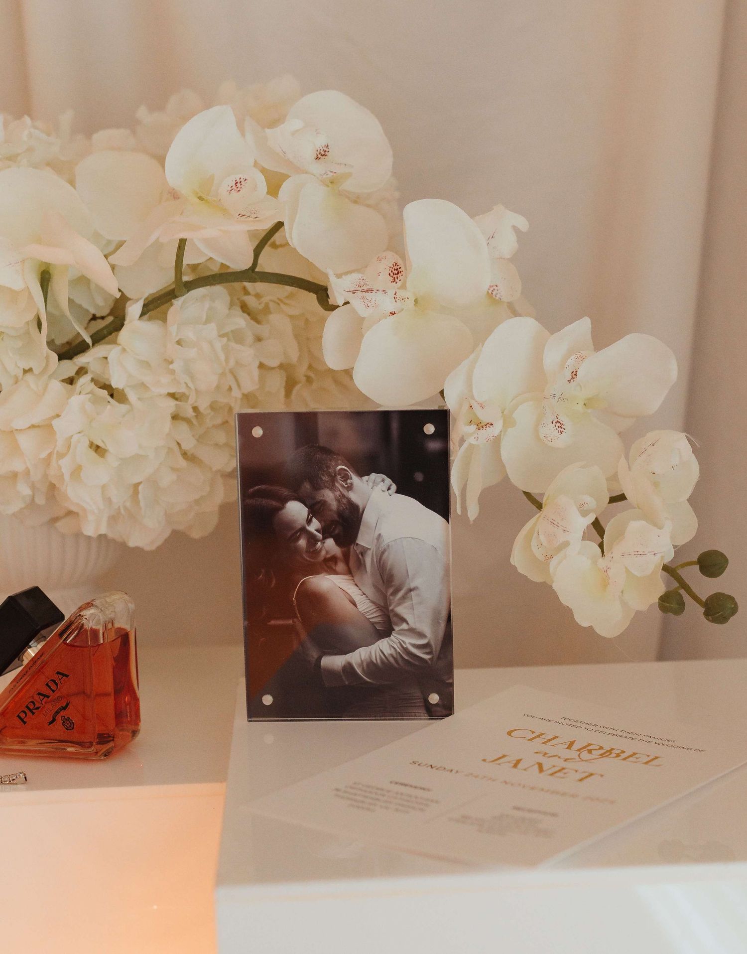 White orchids and hydrangeas arranged beside a framed photo and perfume bottle on a light surface.