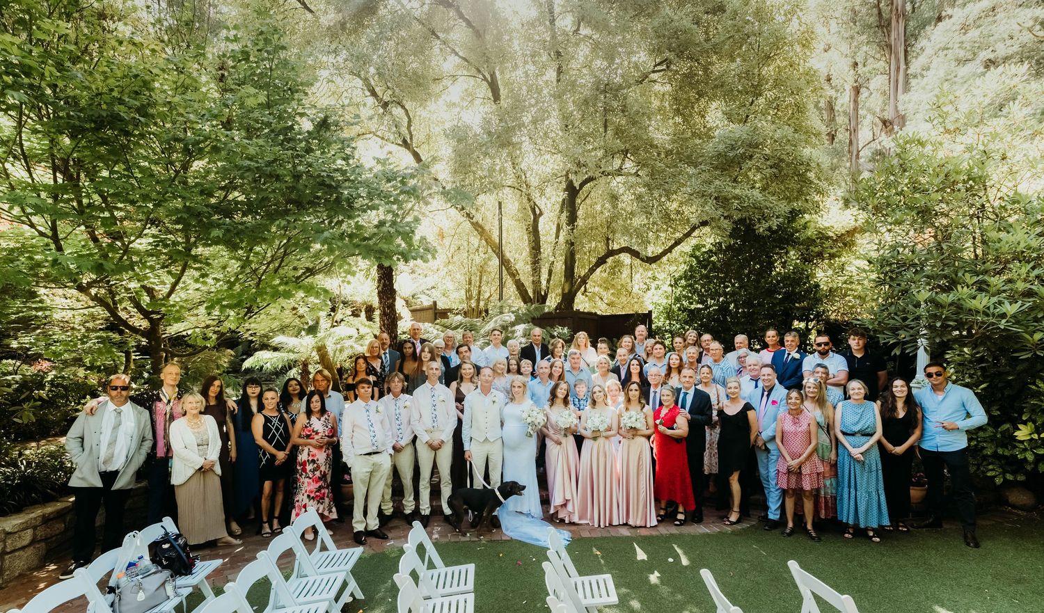 A large outdoor wedding party gathers for a group photo under shady trees in a garden setting.