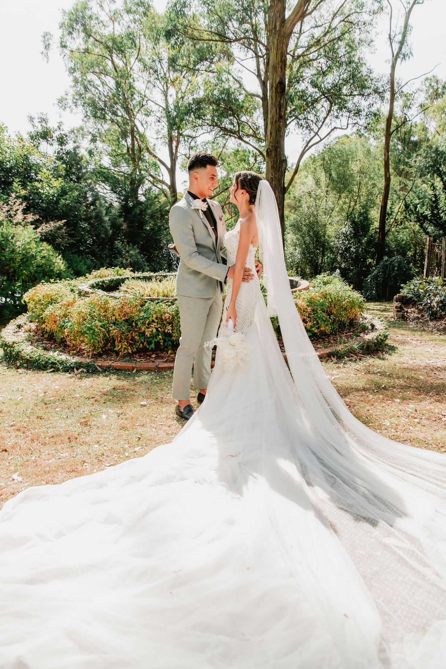 Wedding couple embraces in garden setting with dramatic flowing white veil and grey suit against lush greenery backdrop.