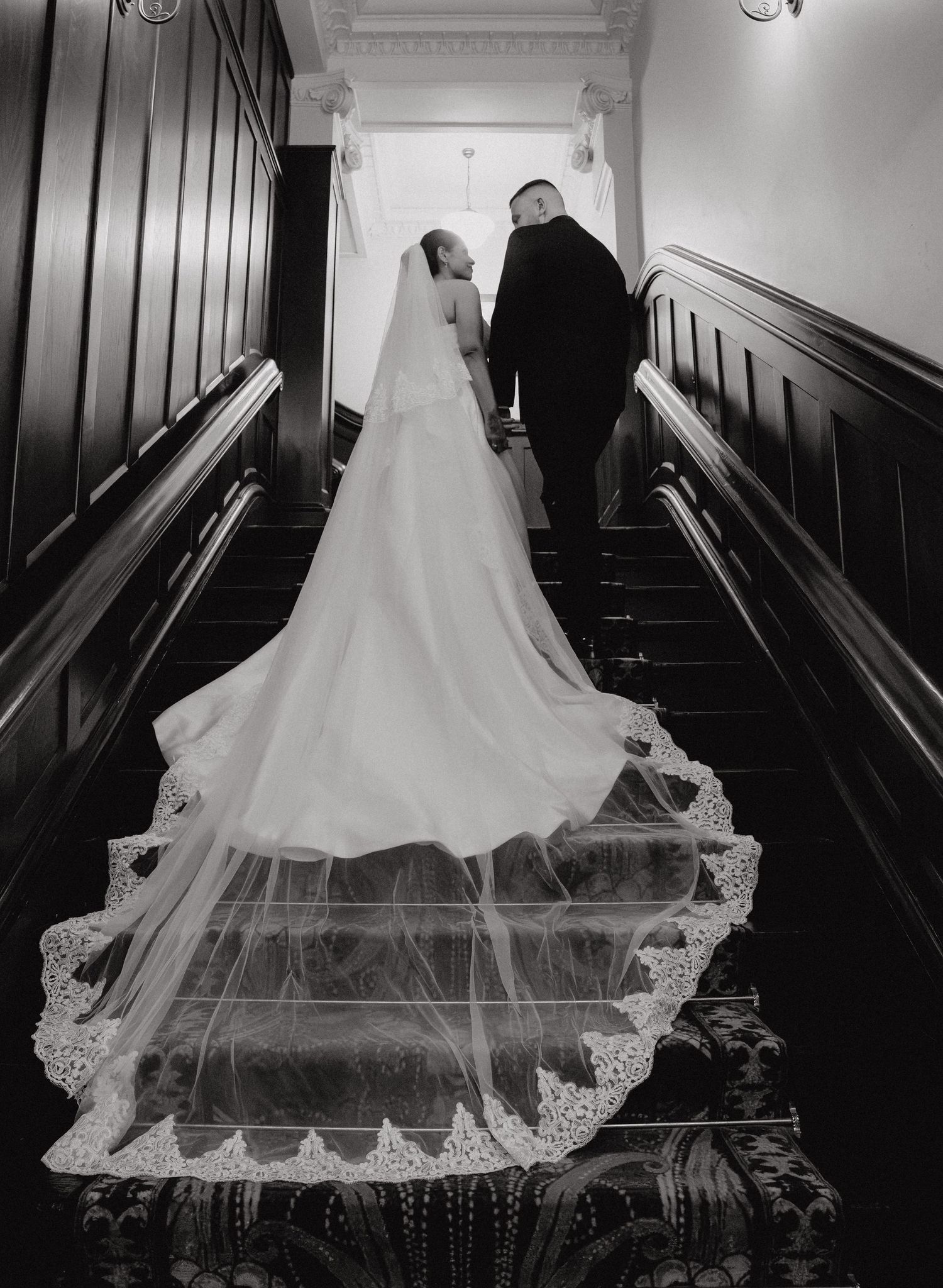 Black and white photo of bride and groom ascending grand staircase, long lace cathedral veil trailing elegantly behind.
