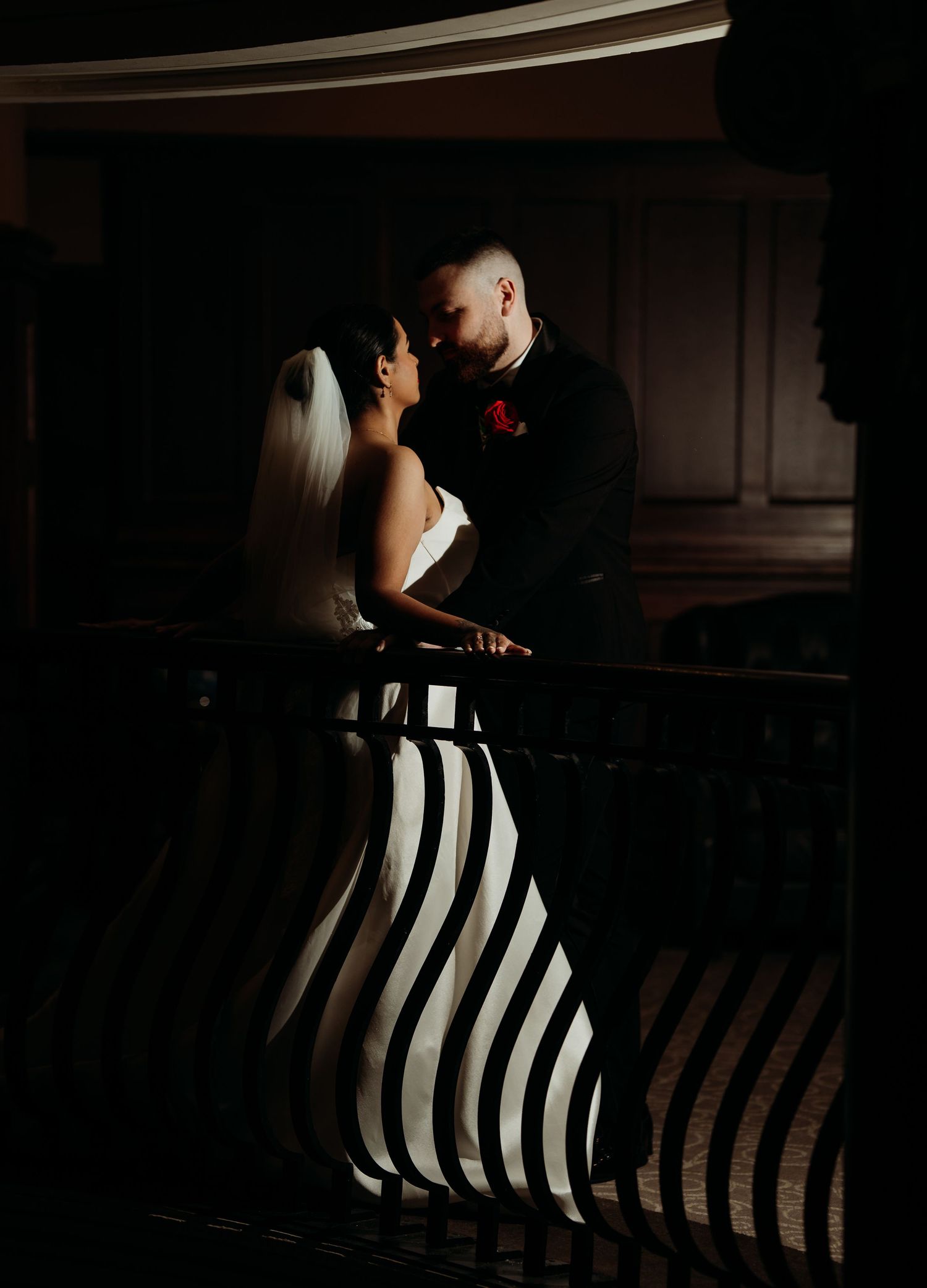 Bride in bold black and white striped gown shares romantic moment with groom in black suit on dimly lit balcony.