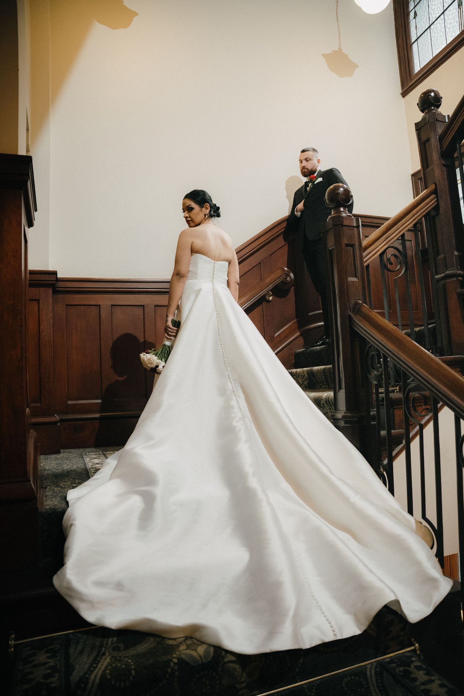 Bride in elegant white ball gown with long train poses on wooden staircase with groom in black suit behind her.