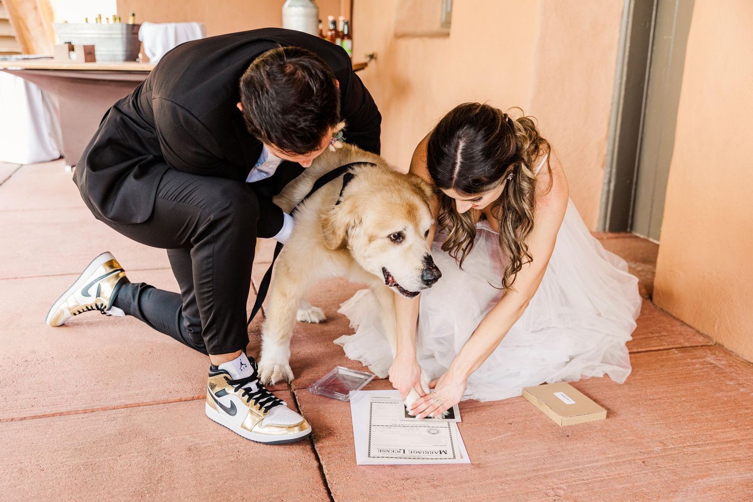 A couple in wedding attire kneels on the floor with their golden retriever while signing a document together.