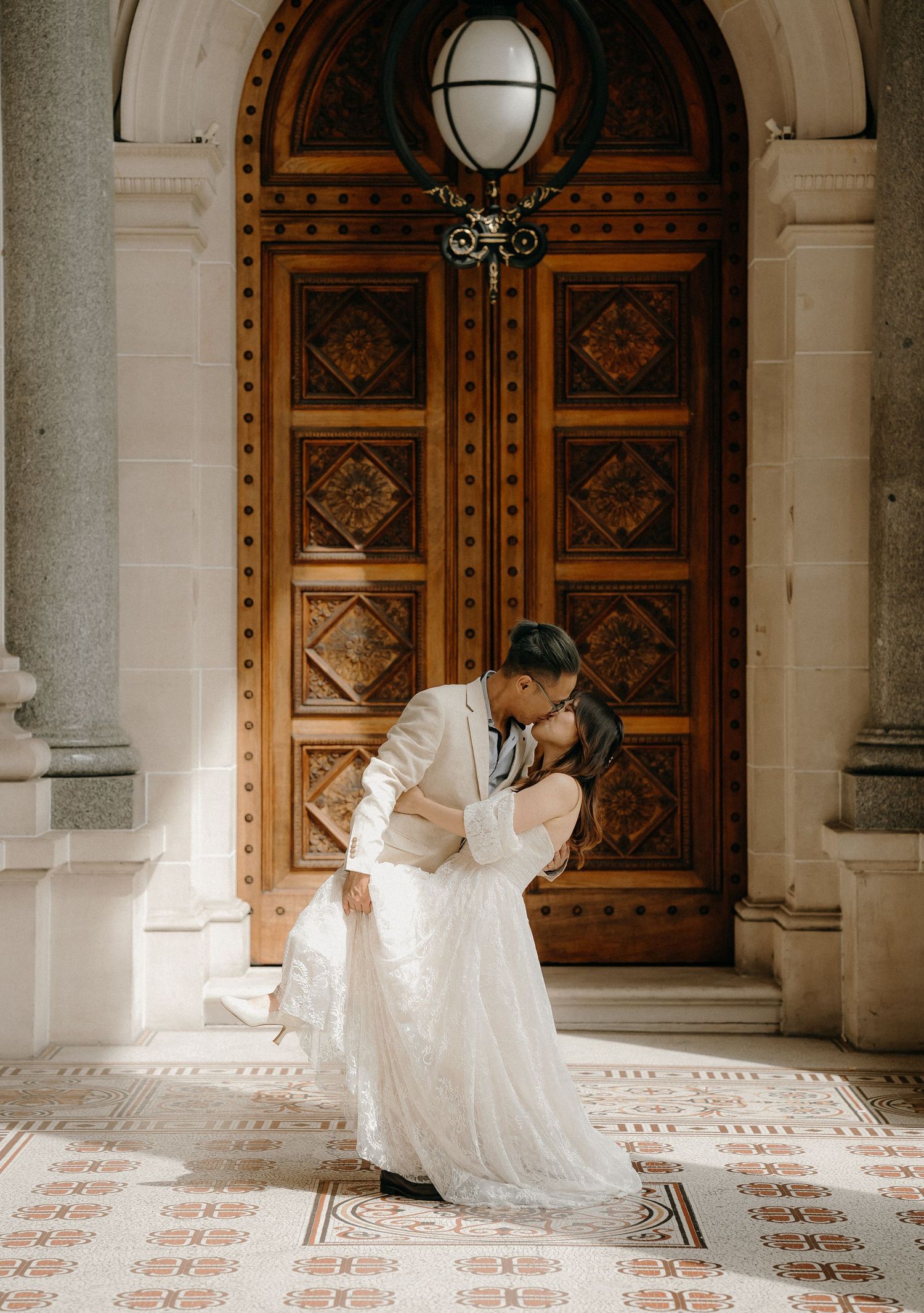 A romantic couple in wedding attire share an intimate moment in front of an ornate wooden door with stone columns.