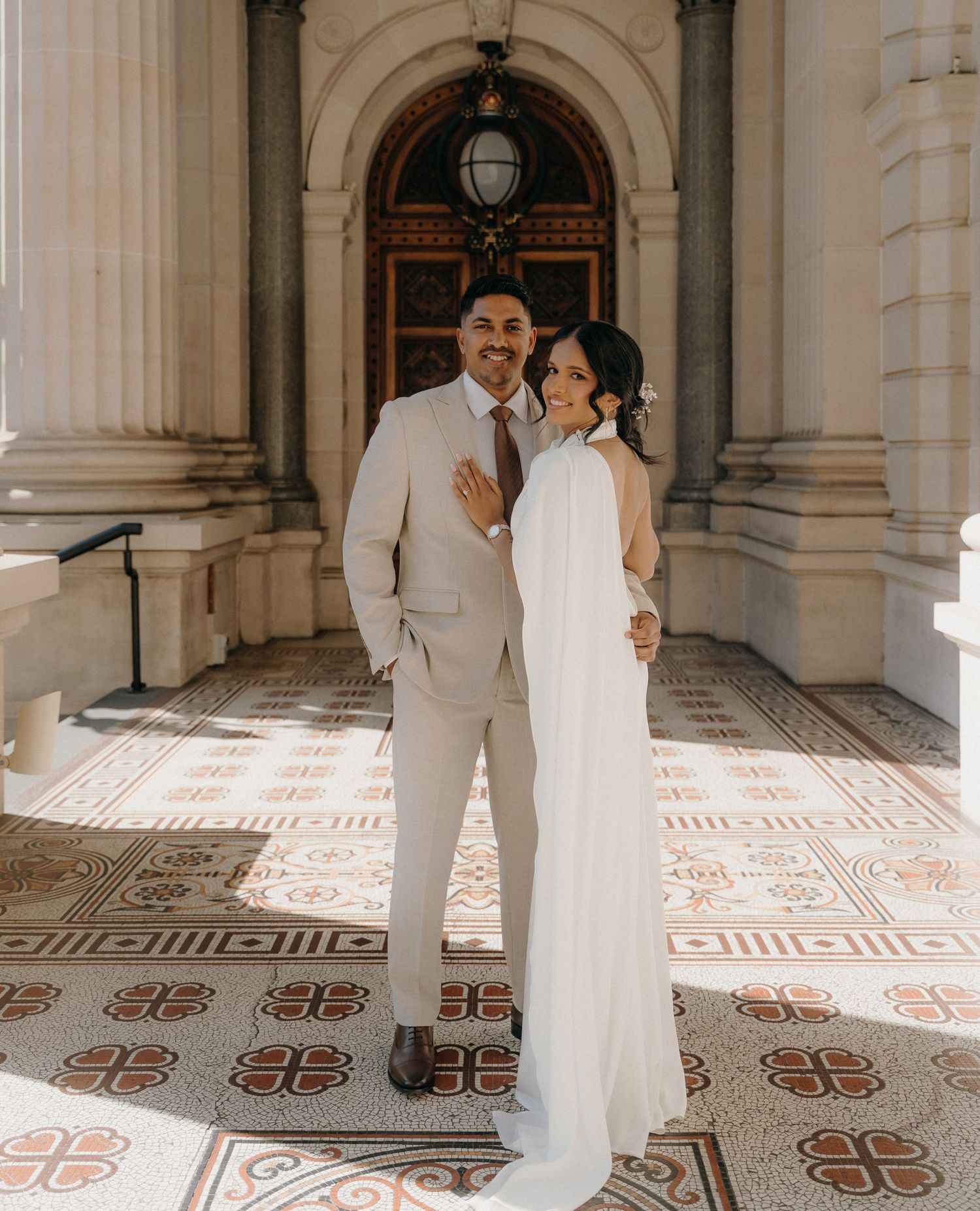 Couple in elegant wedding attire poses in a grand hallway with ornate tiled floors and classical architecture.