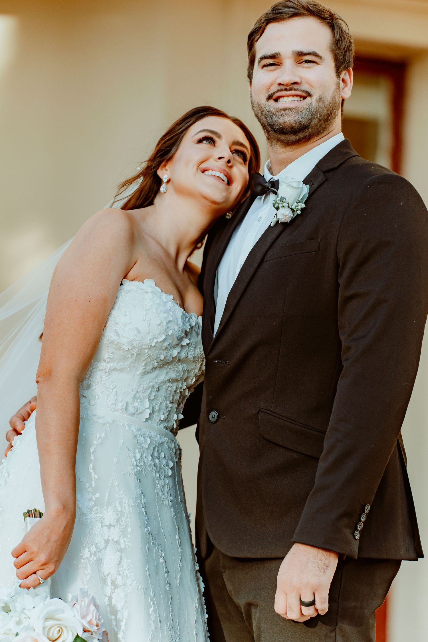 A happy newlywed couple in wedding attire embracing outdoors, with the bride in a beaded white dress and groom in dark suit.