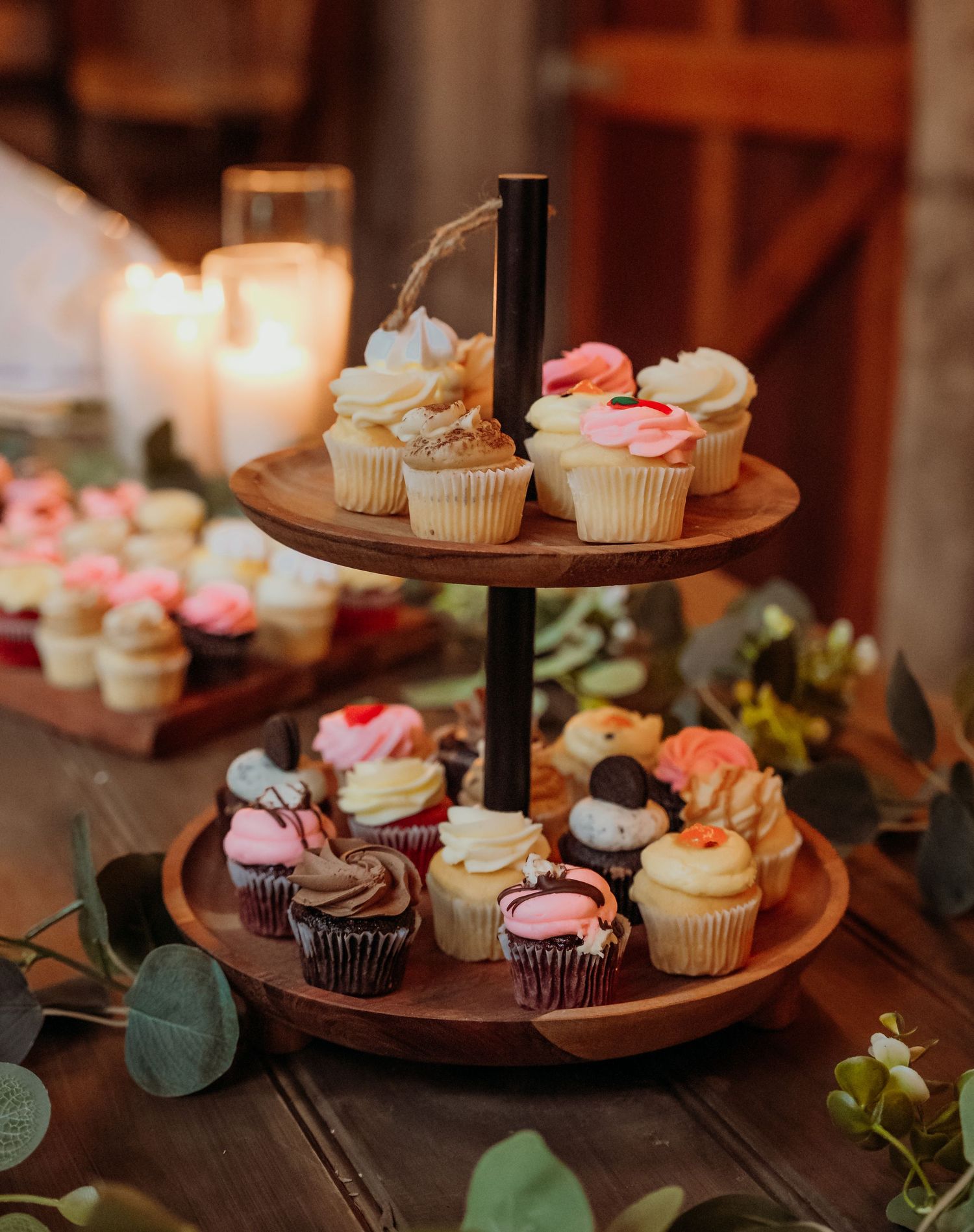 Two-tiered wooden dessert stand displaying an assortment of decorated cupcakes with pink and white frosting and greenery.