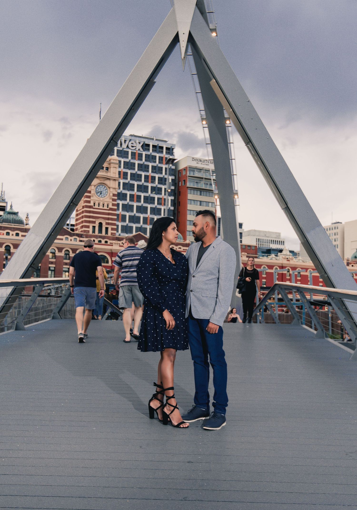 A couple shares a romantic moment on a modern bridge with geometric architecture and cloudy skies in the background.