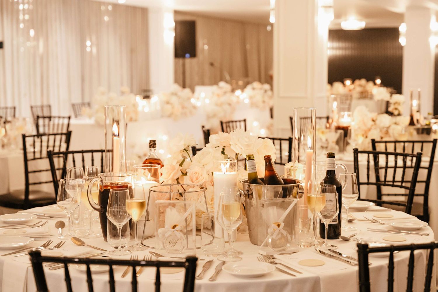 Elegant wedding reception hall with white table settings, floral centerpieces, and illuminated black chiavari chairs.