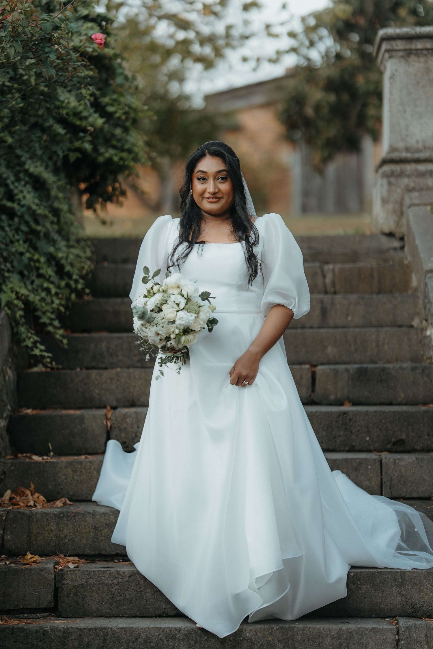 A bride wearing a flowing white gown with puff sleeves holds a white bouquet while standing on stone steps outdoors.
