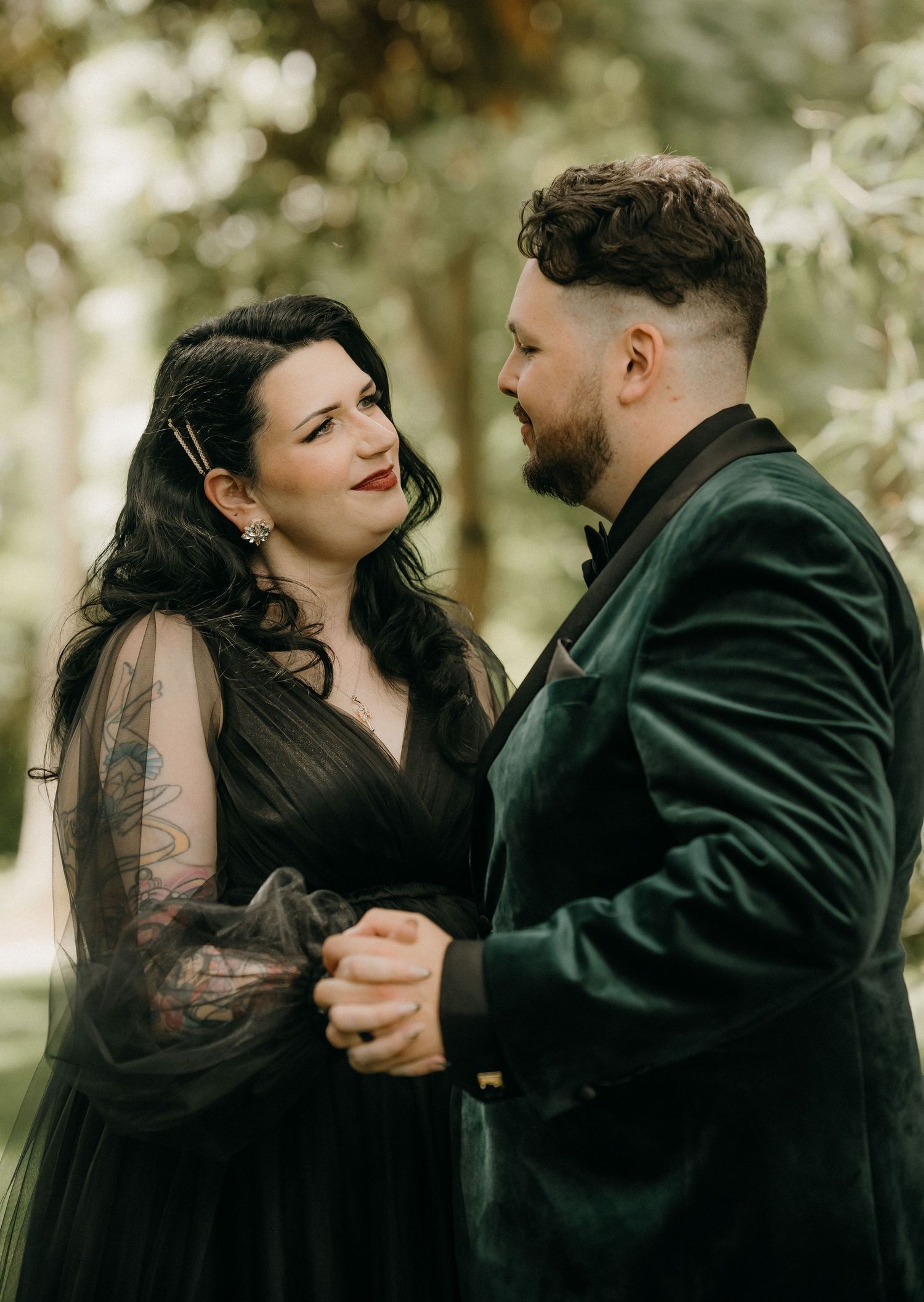 A couple in dark formal attire share an intimate dance moment in a dreamy outdoor setting.