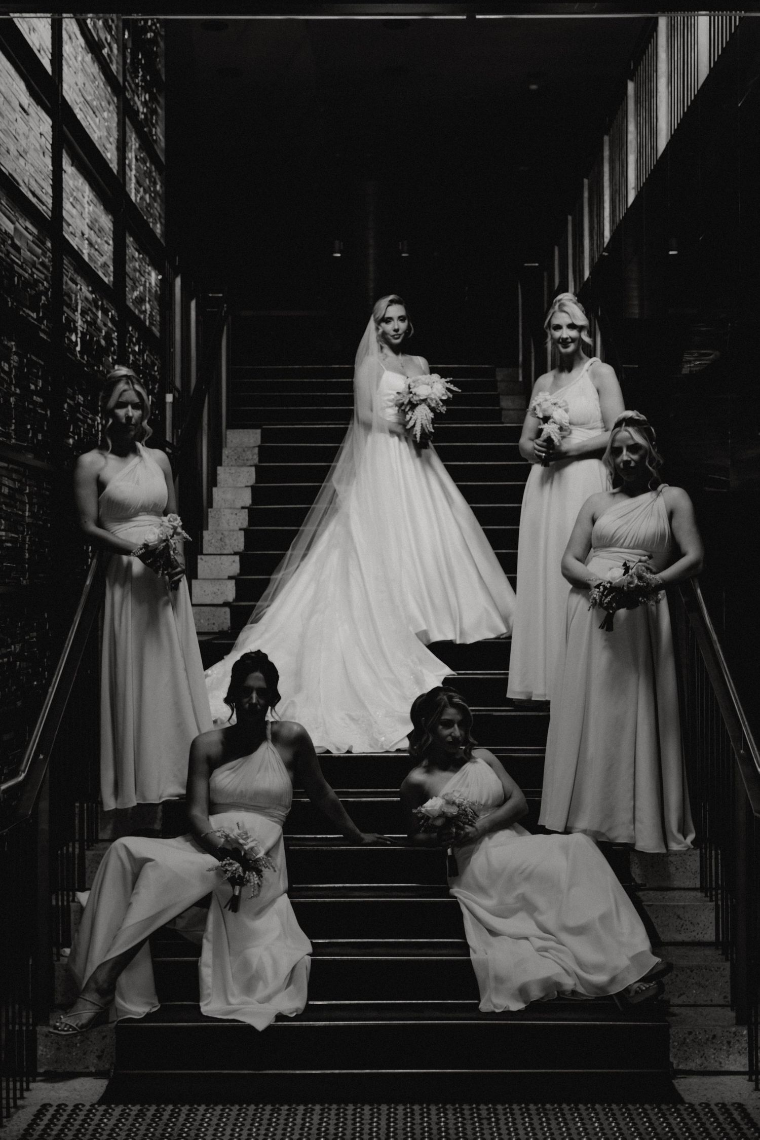 A black and white wedding photo showing a bridal party elegantly posed on stairs in flowing light-colored gowns.