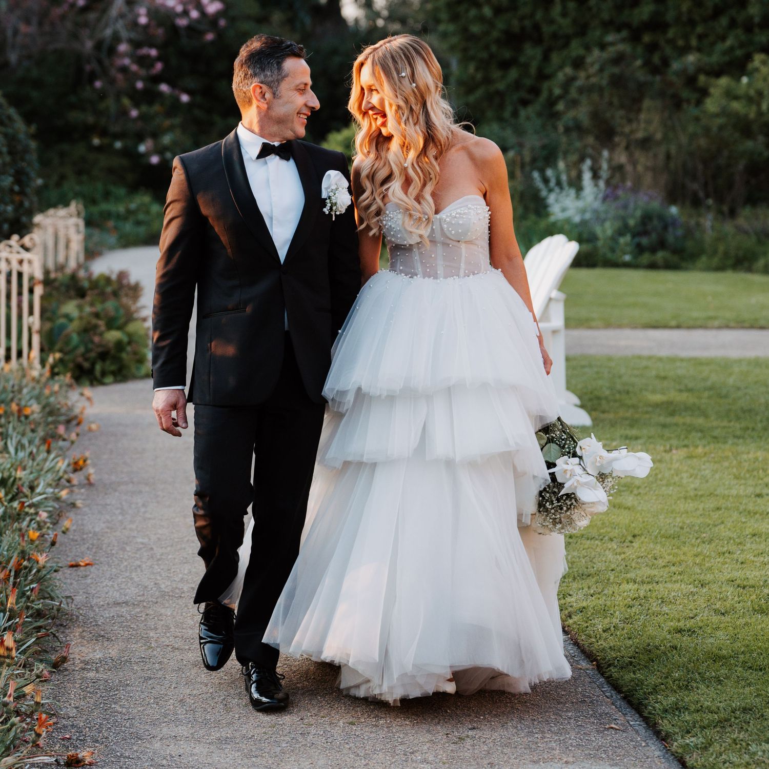 A romantic couple walks along a garden path in formal wedding attire during a golden sunset.
